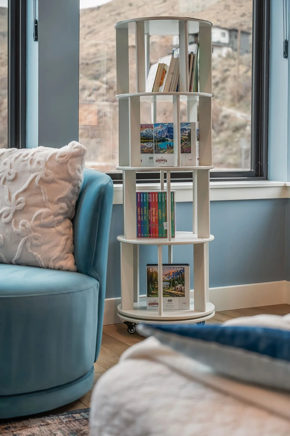 White circular bookshelf with tiered shelves on casters, holding books and puzzles, positioned near a window with a mountain landscape outside, beside a blue armchair with embroidered pillow.