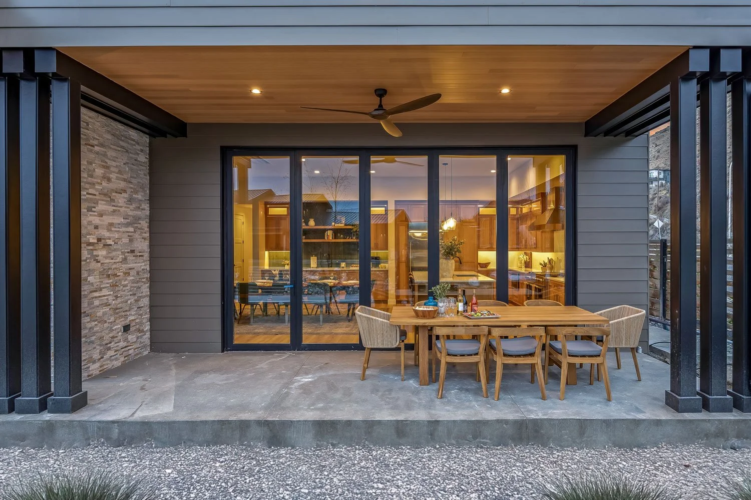 Outdoor patio area with wooden dining table and chairs, covered by a ceiling with a ceiling fan. Behind a glass sliding door, a well-lit kitchen and dining space are visible inside the house.