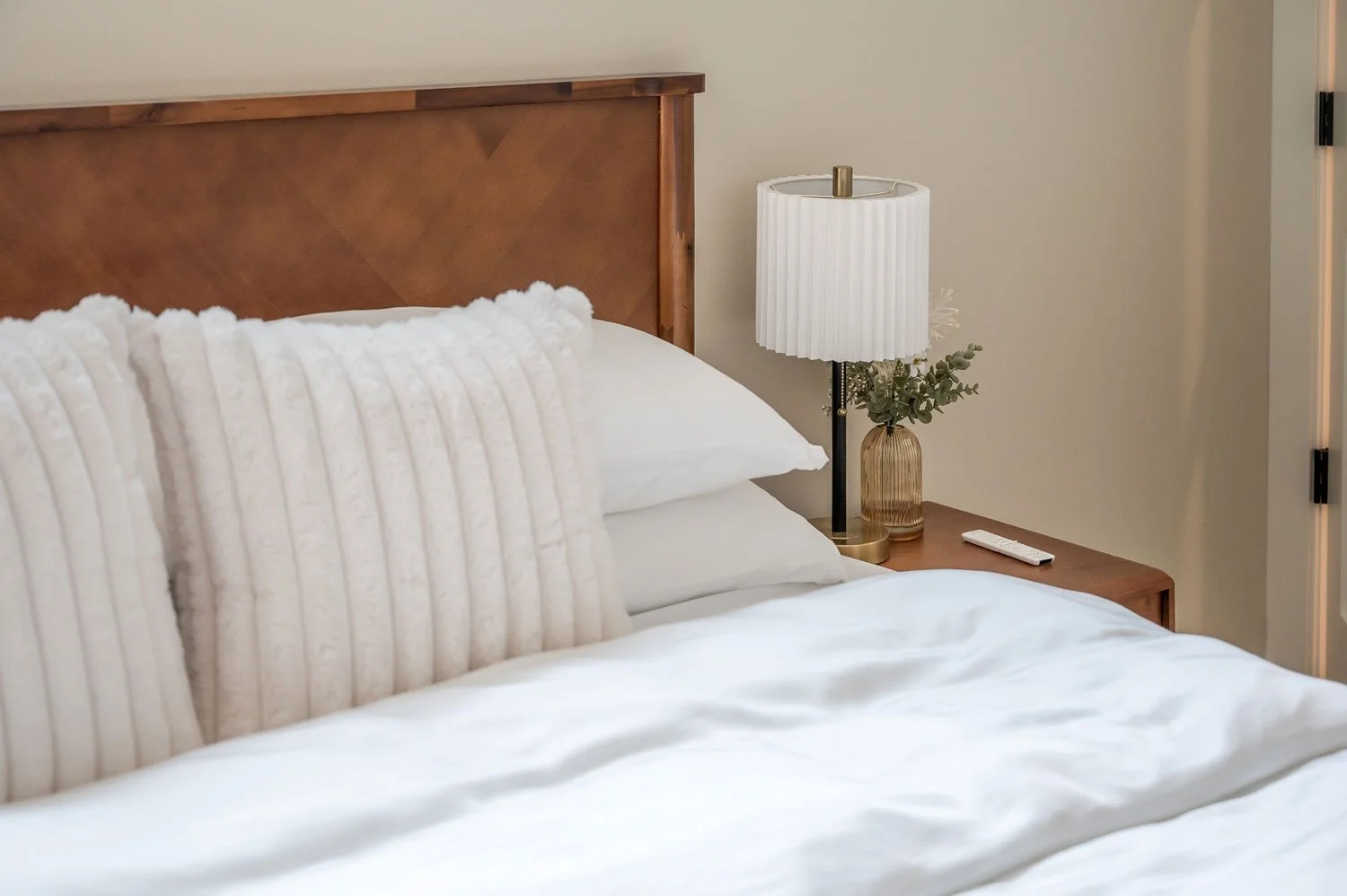 A neatly made bed with white pillows and duvet, a wooden headboard, a bedside table with a white lamp, a vase with greenery, and a remote control.