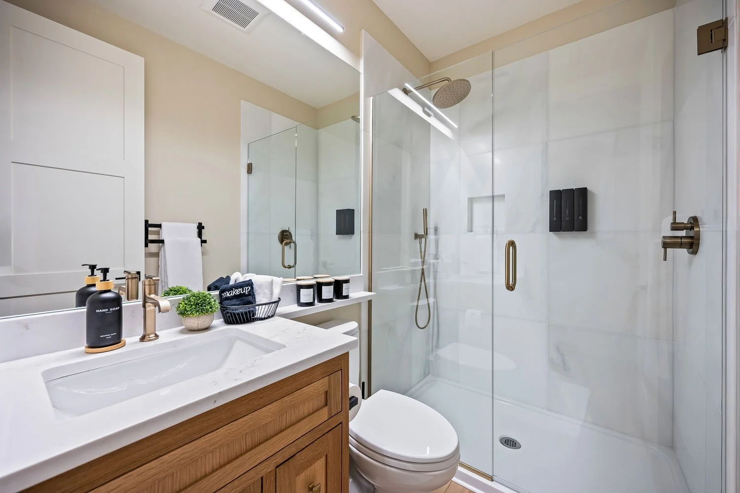 Modern bathroom with glass shower enclosure, wooden vanity, white marble countertop, and minimal decor including hand soap, hand towel, small plants, and black candles.