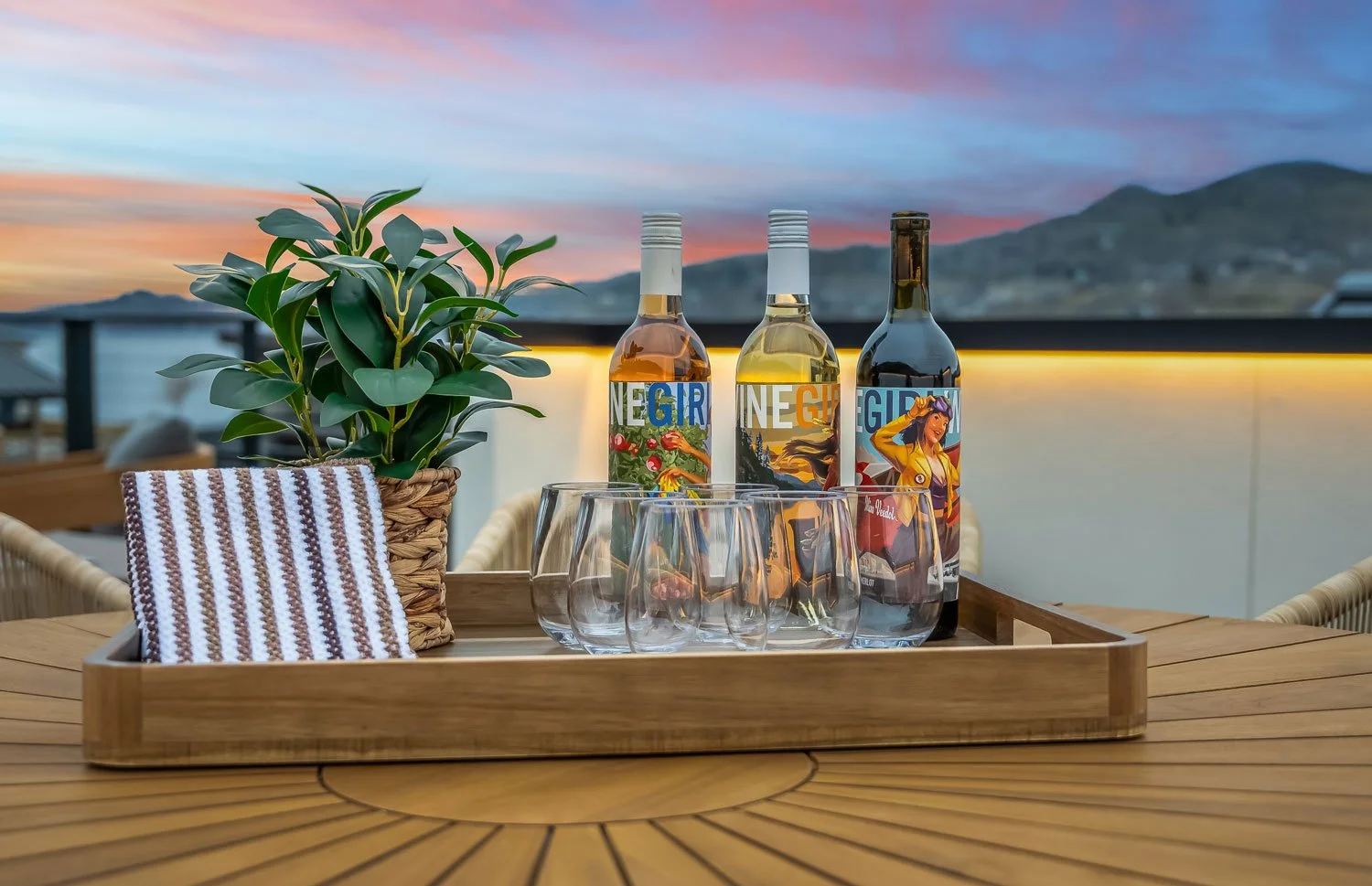 A wooden table with a tray holding three bottles of wine, four empty glasses, a potted plant, and a striped cloth, set against a sunset sky and mountains in the background.