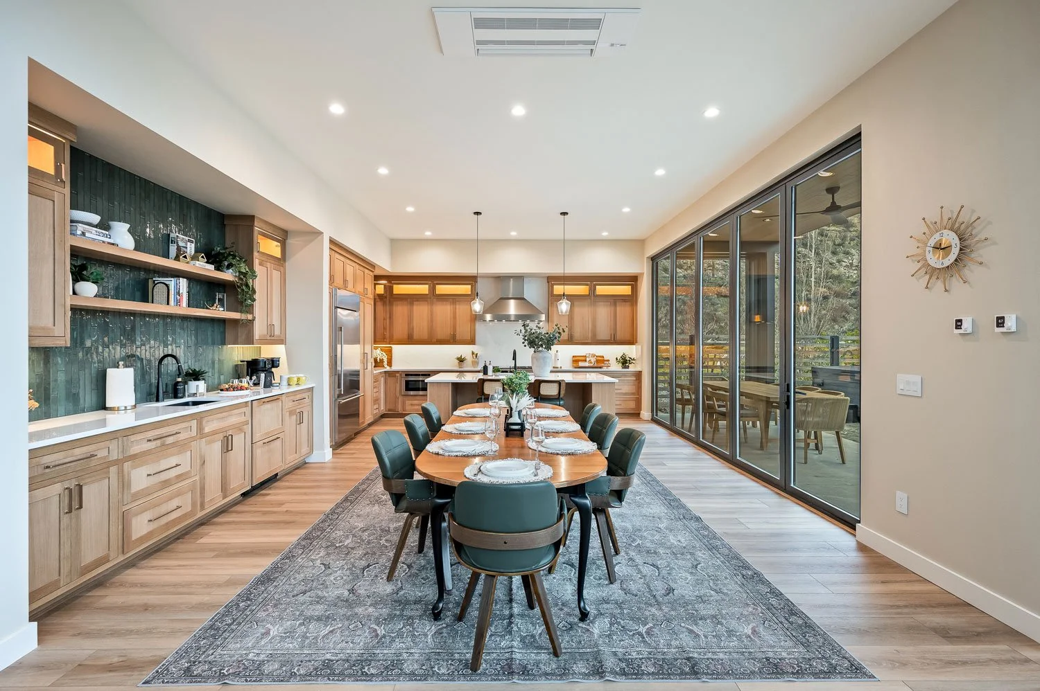 Modern dining room and kitchen with wooden cabinetry, a large wooden table set with plates and glasses on a gray patterned rug, and large sliding glass doors leading to an outdoor patio with trees in the background.