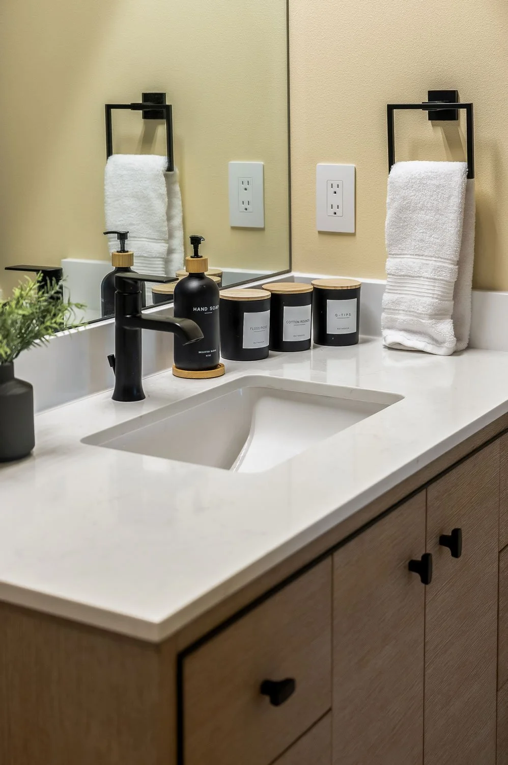 Bathroom sink with black soap dispenser, two black jars labeled 'Floss', 'Cotton Rounds', and 'Q-Tips', a white hand towel hanging on a black towel holder, and a small plant on the left side.
