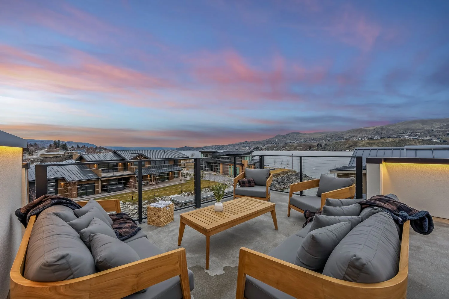 Outdoor rooftop patio with wooden furniture, gray cushions, and a view of neighboring modern houses and rolling hills at sunset.