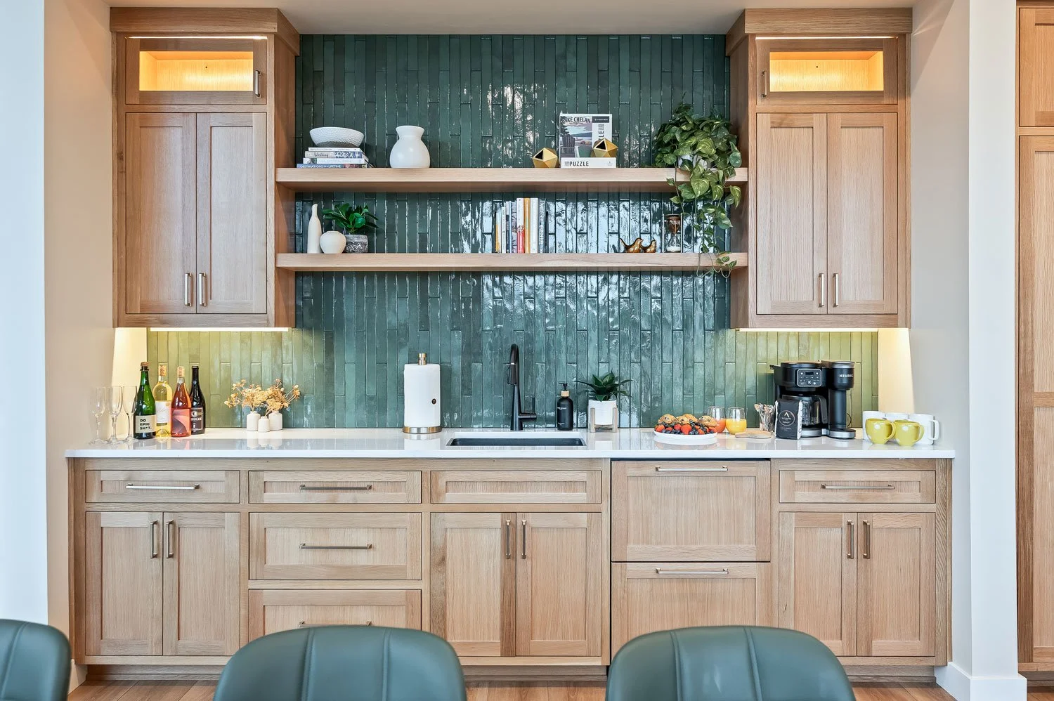 Modern kitchen with light wooden cabinets, green tiled backsplash, open shelves with decorative items, kitchen counter with coffee maker, bowls, and drinks, and green chairs in the foreground.