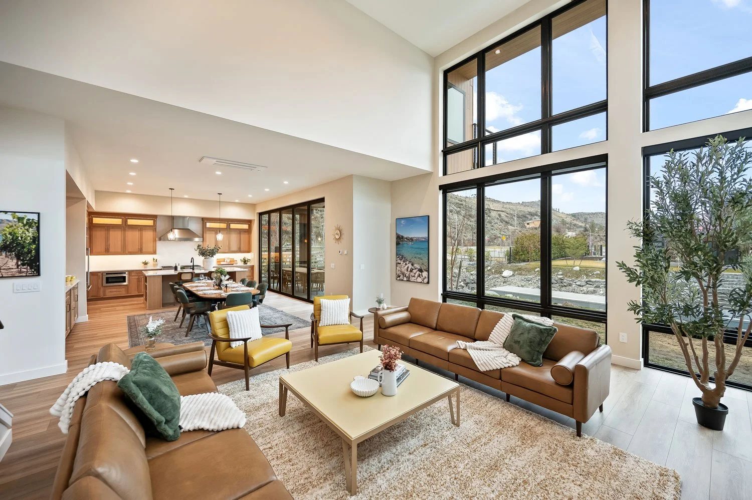 Open-concept living room with large floor-to-ceiling windows overlooking mountains, tan sofas, green and yellow accent chairs, a white coffee table with decor, and a view of a dining area and modern kitchen with wooden cabinets.