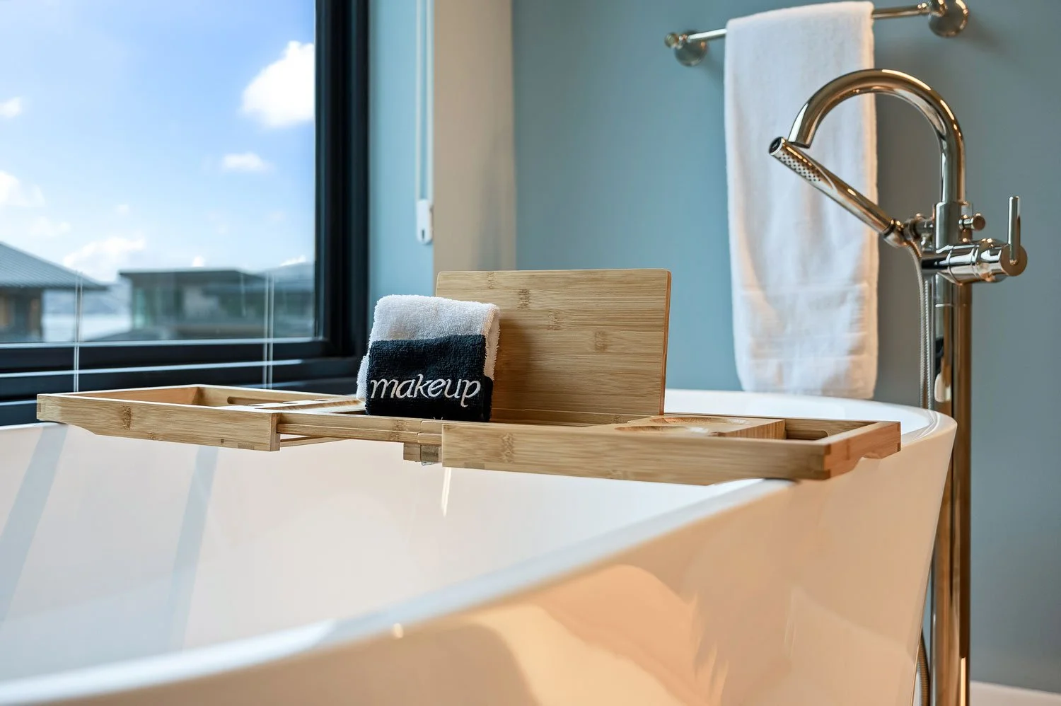A cozy bathroom with a white bathtub, a wooden bath tray holding a black and white towel with the word 'makeup' embroidered on it, and a window showing an outdoor view of roofs and a blue sky.
