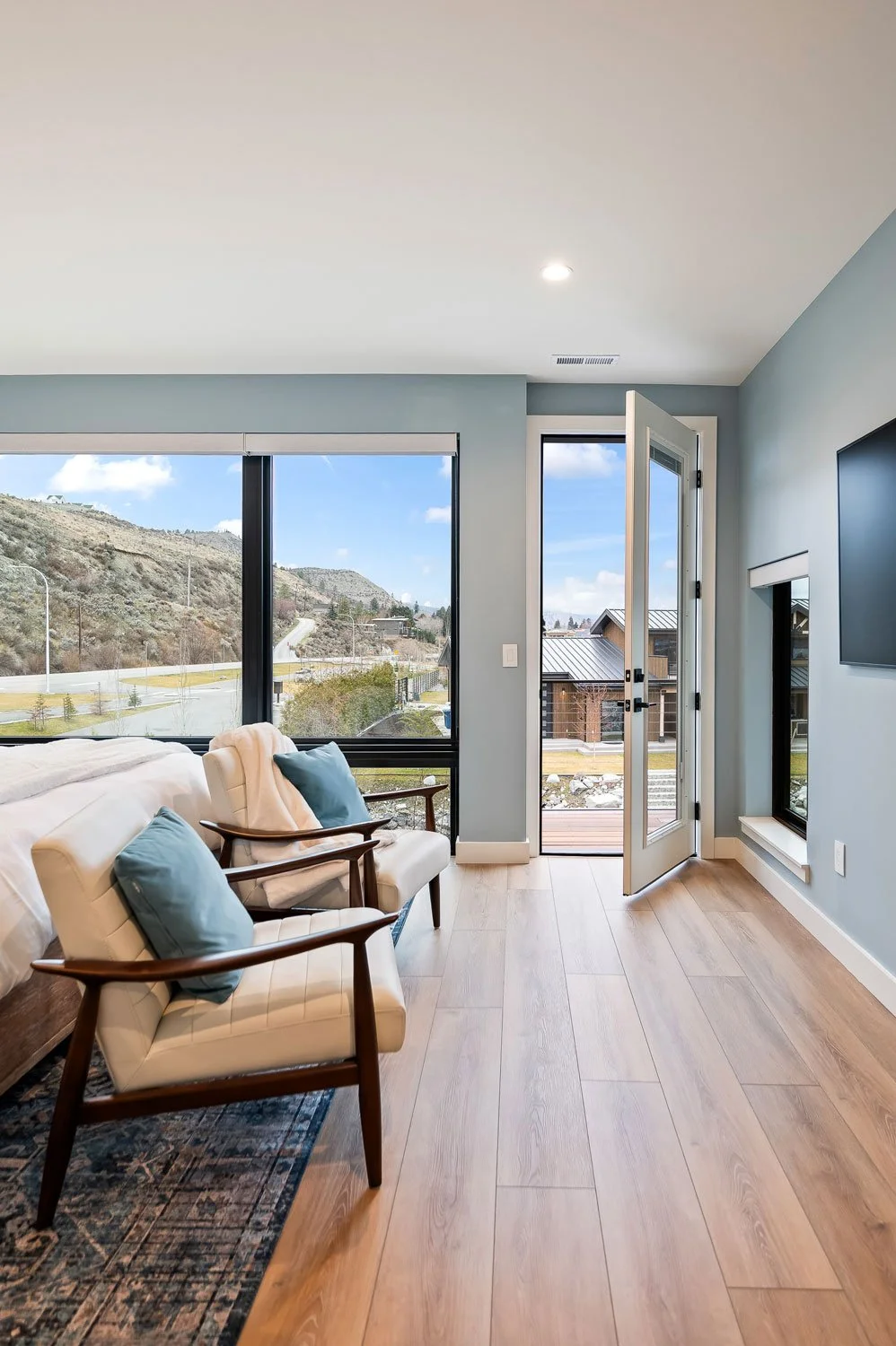Modern living room with light wood flooring, large windows showing a hillside view, glass door leading to a balcony, and a wall-mounted TV.