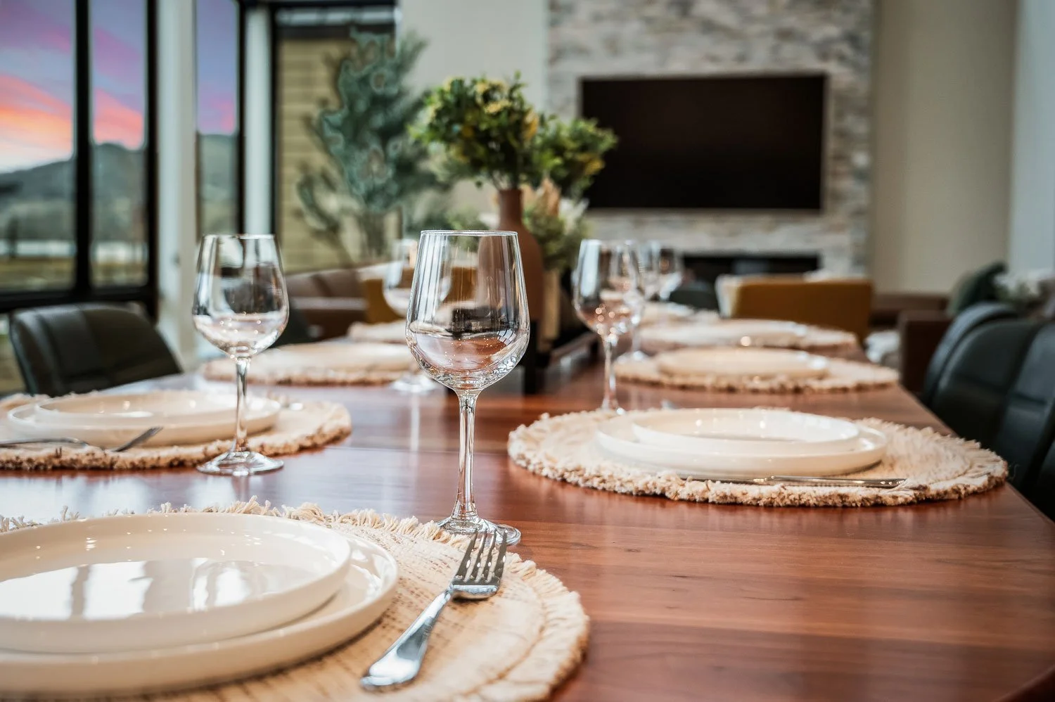 Empty table set with white plates, silverware, and empty wine glasses, in a dining room with large windows and a fireplace.