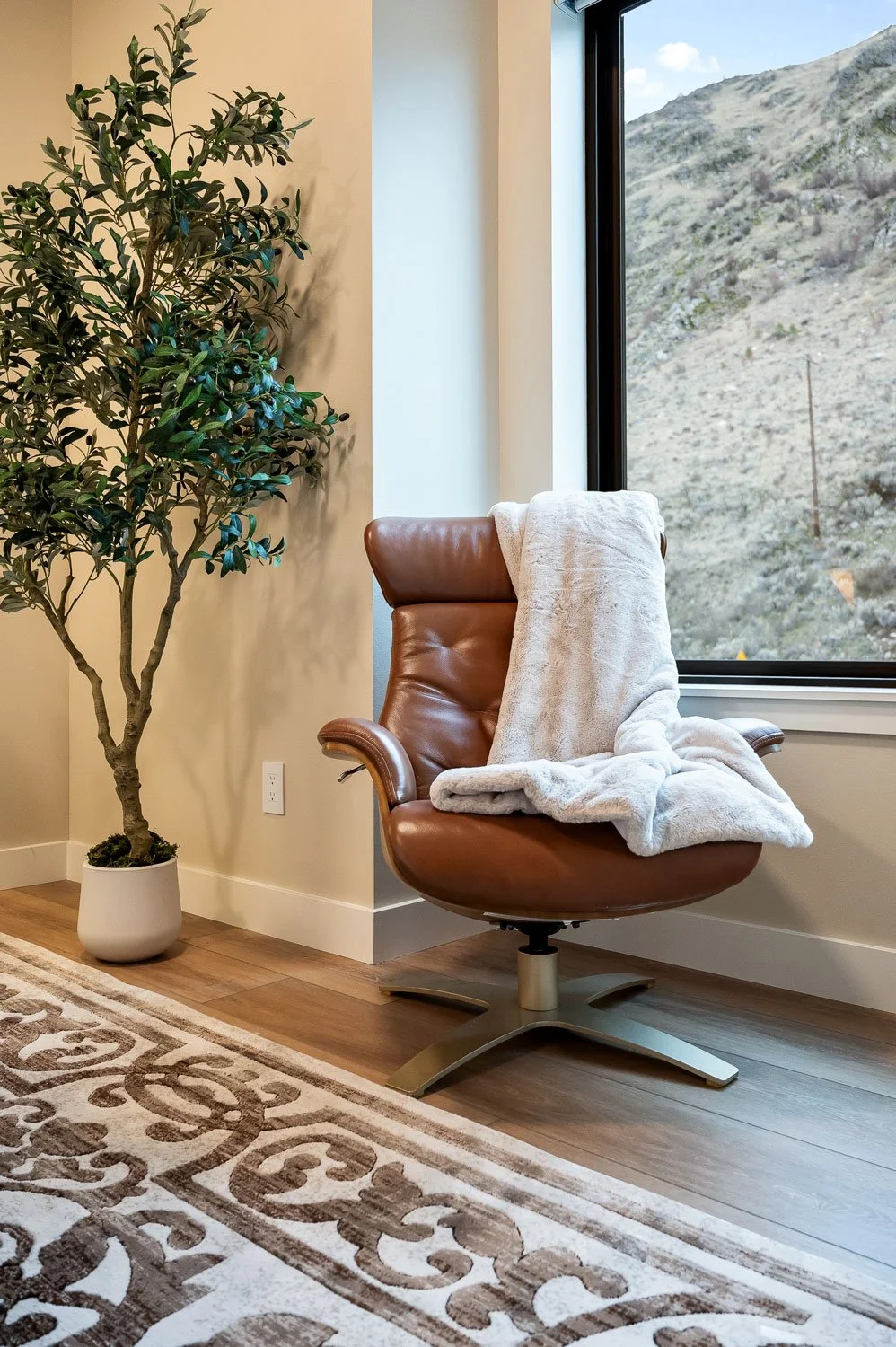 A cozy corner with a brown leather swivel chair draped with a plush white blanket next to a large window showing a mountainous landscape, and a tall green potted plant on a wooden floor with a patterned area rug.