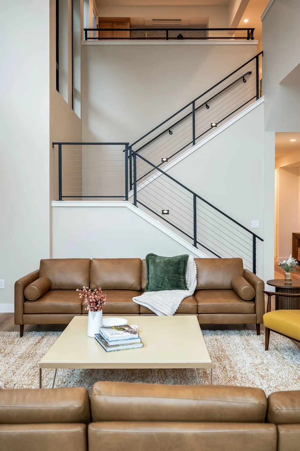 Living room with a leather sofa, cozy blankets and pillows, and a coffee table with books and flowers. A staircase with black and white railings leads to an upper level.