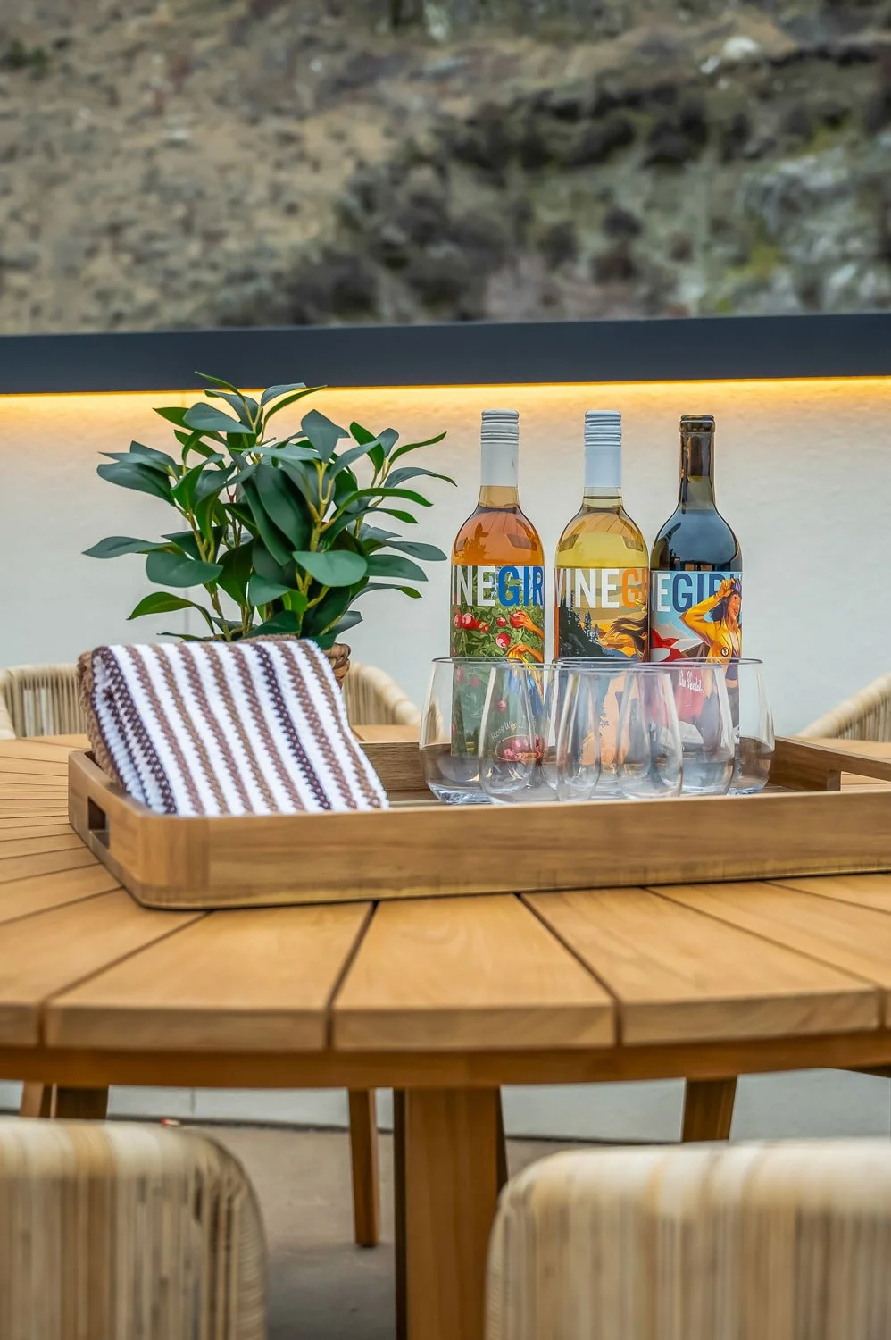 Table with three bottles of flavored soda water, a green potted plant, a wooden tray with a striped cloth, and clear glasses, set against a neutral indoor background with mountains visible outside.