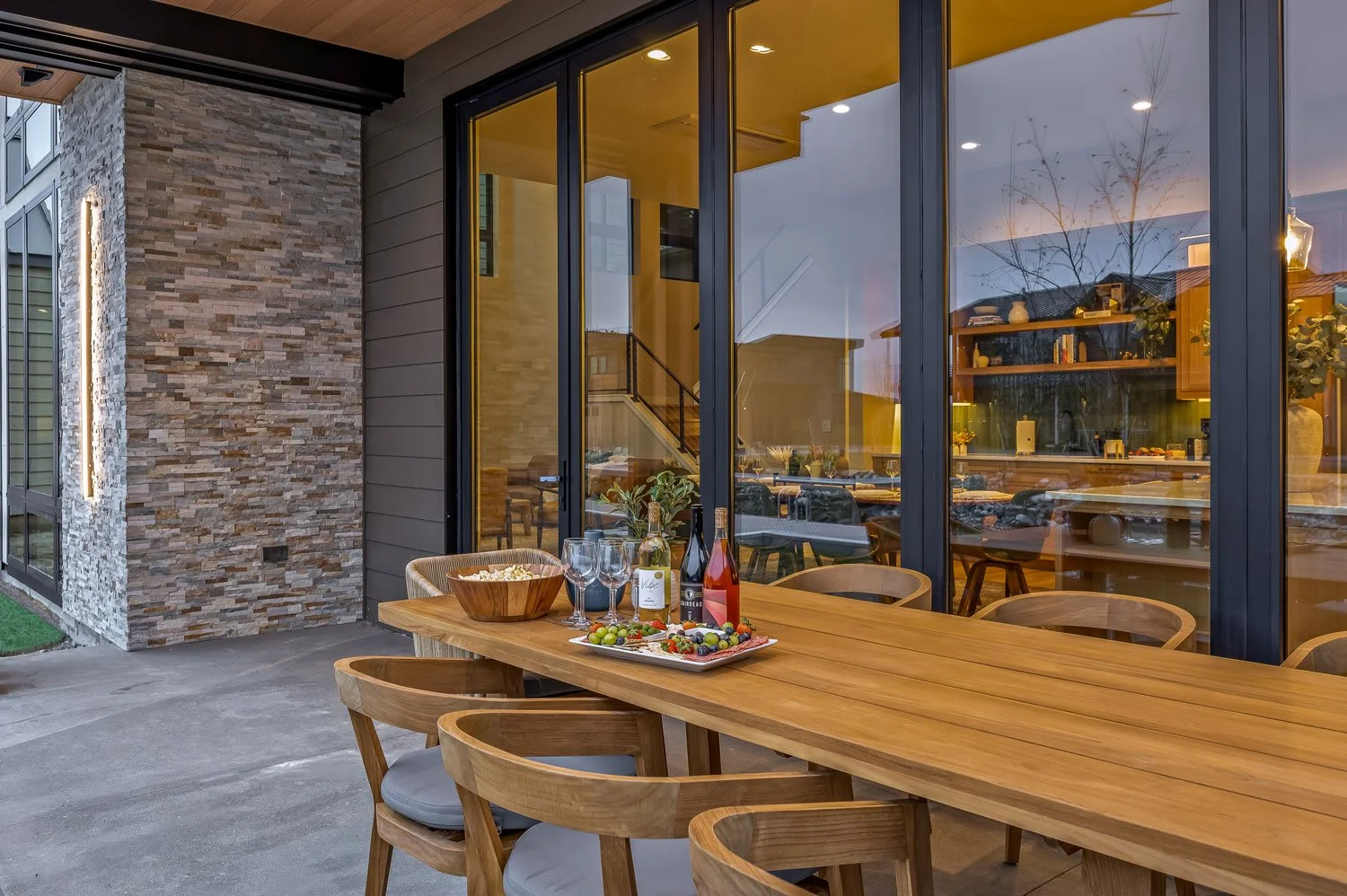 An outdoor dining area in front of a modern house with large glass windows, showing a wooden table set with wine bottles, glasses, a bowl of chips, and a fruit platter, illuminated softly at dusk.