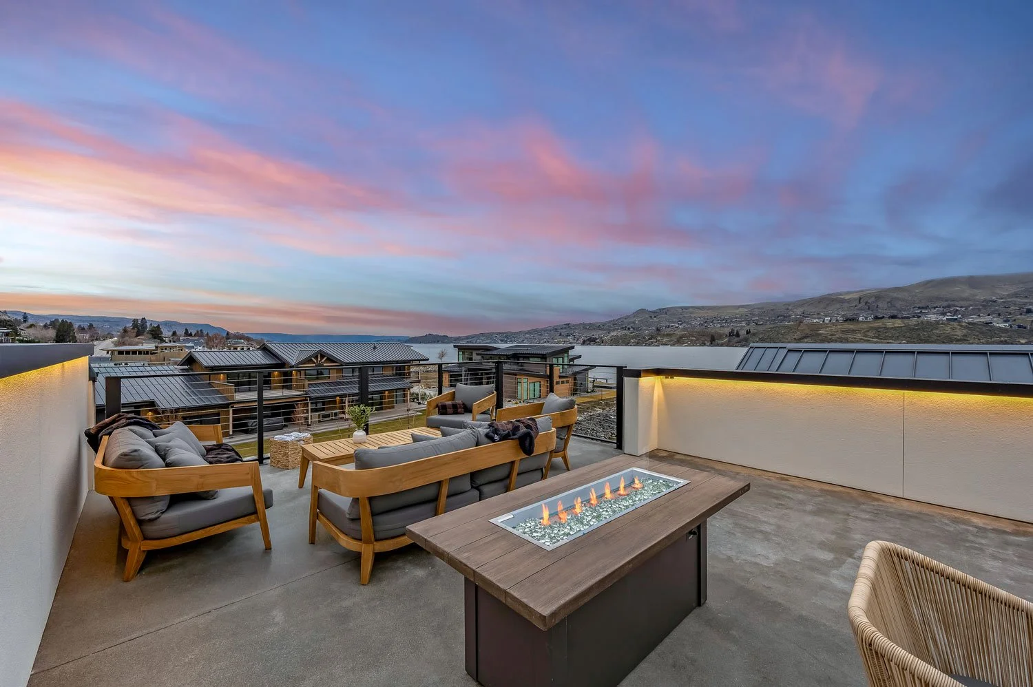 Rooftop patio with wooden seating, a rectangular fire pit table, and scenic view of hills and water during sunset.