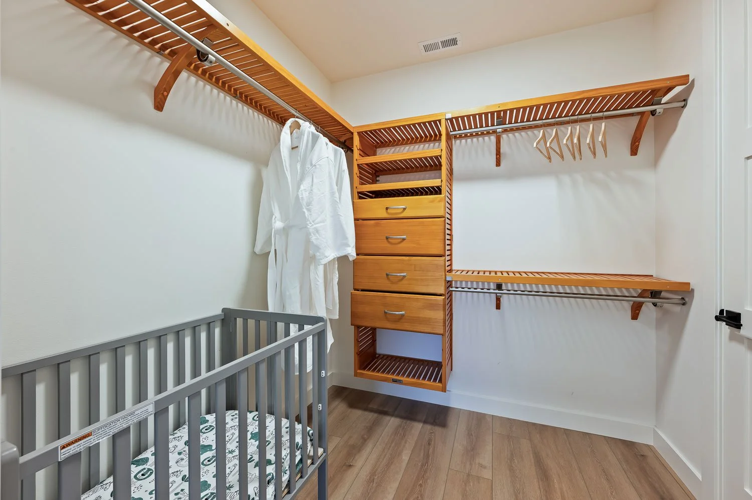 Empty walk-in closet with wooden shelves, drawers, and hanging rods, a gray crib with a patterned mattress, and a white bathrobe hanging in the closet.