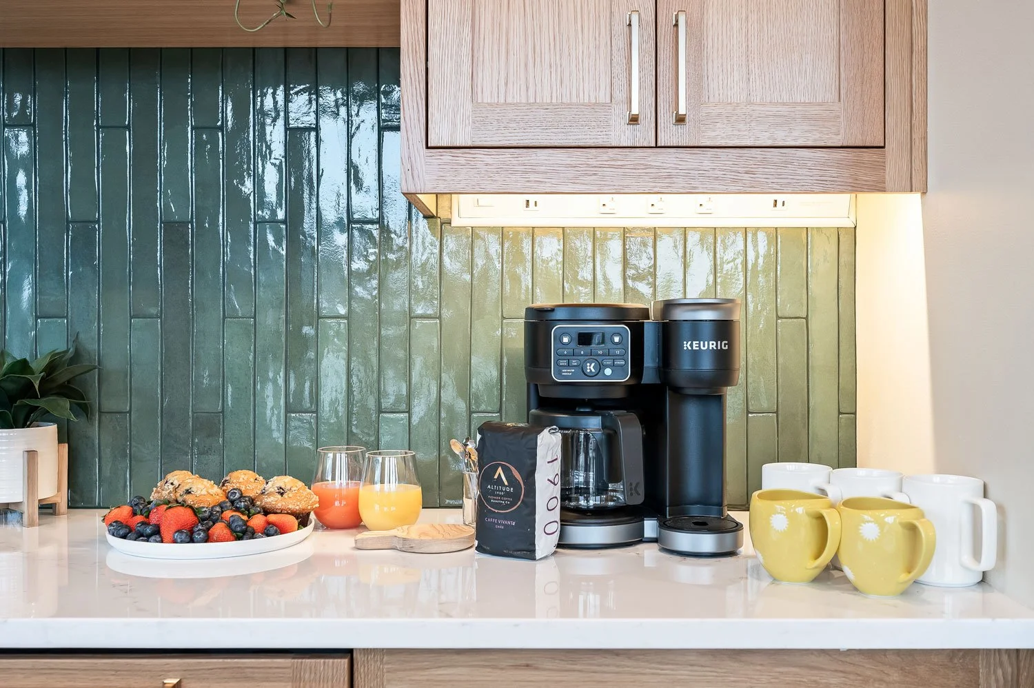 Coffee maker with cups, fruit plate with berries and muffins, glasses of orange juice and pink beverage on kitchen counter with green backsplash and wooden cabinets.