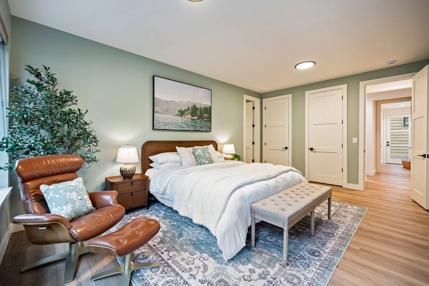 A bedroom with a large bed, white bedding, and pillows, flanked by nightstands with lamps, a brown leather lounge chair with a pillow, a large potted plant, and a framed lake landscape picture on the wall, with parquet flooring and multiple closed do