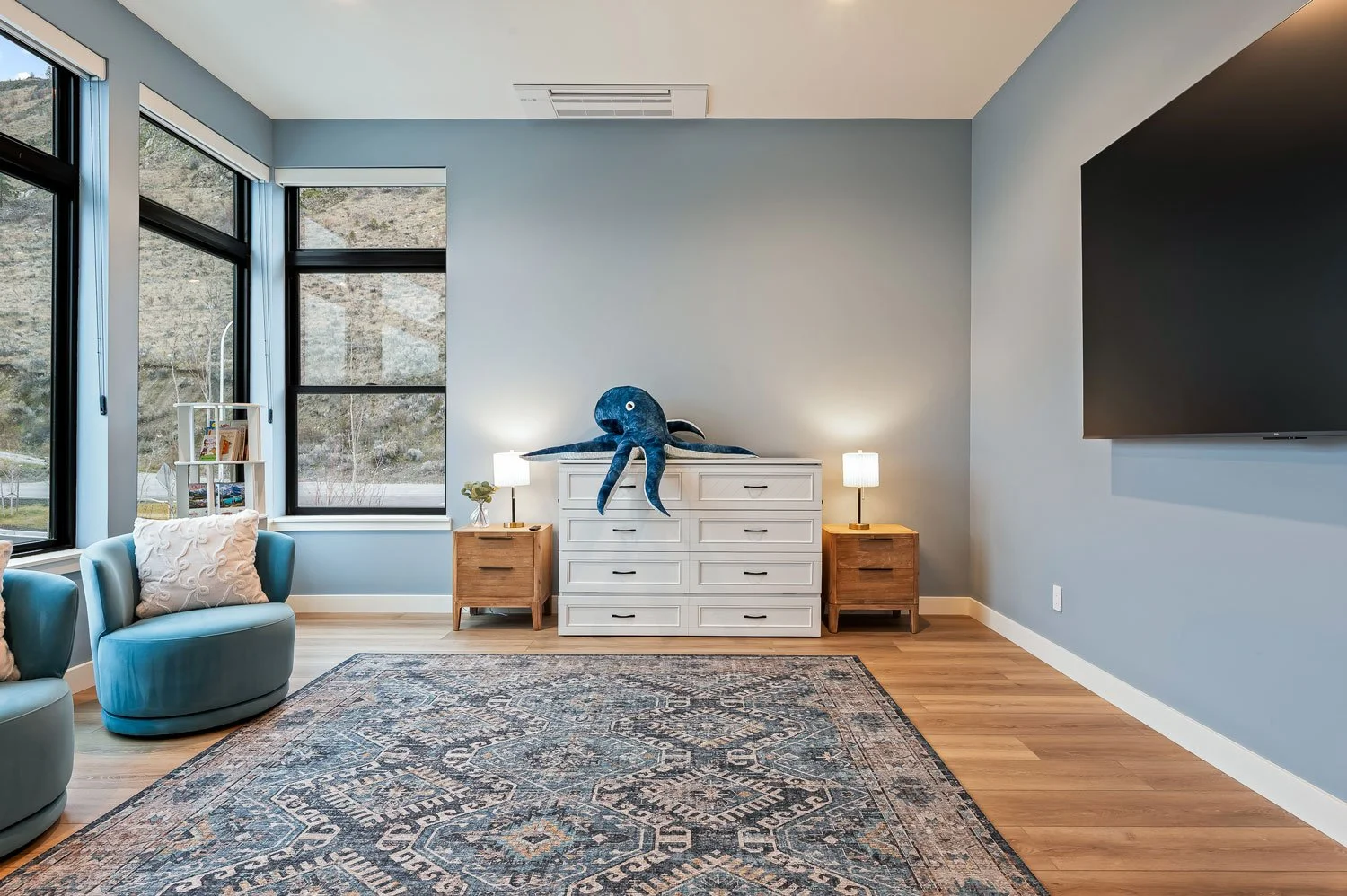 Living room with light blue walls, large windows, two teal armchairs, a white dresser with a blue stuffed octopus on top, two small wooden nightstands with lamps, a patterned rug, and a large black flat-screen TV on the wall.