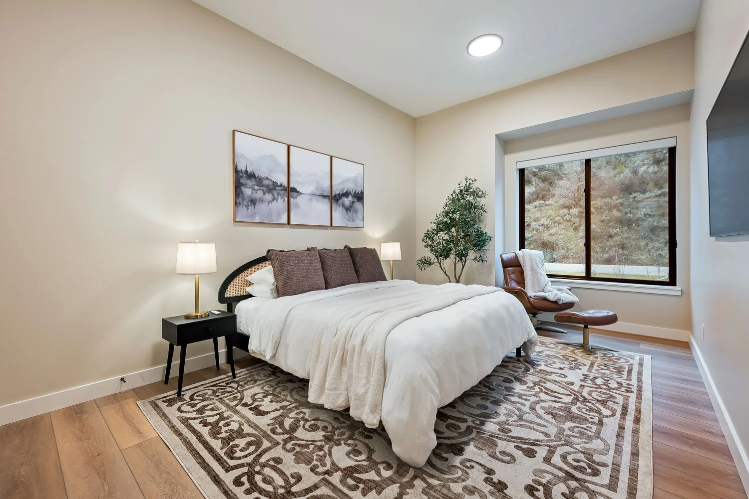A bedroom with a bed, pillows, nightstand with lamp, potted plant, armchair near window, artwork above bed, TV on wall, and wooden flooring.