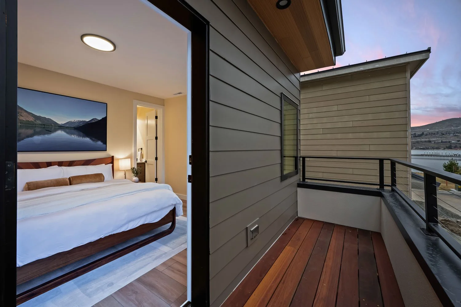 A bedroom with a bed, wall art of a lake and mountains, and a balcony outside with a wooden floor and railing, overlooking a hilly landscape at sunset.