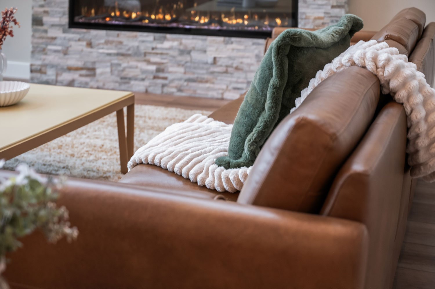 Close-up of a living room couch with green and white throws and pillows, a beige coffee table, and a stone fireplace in the background.