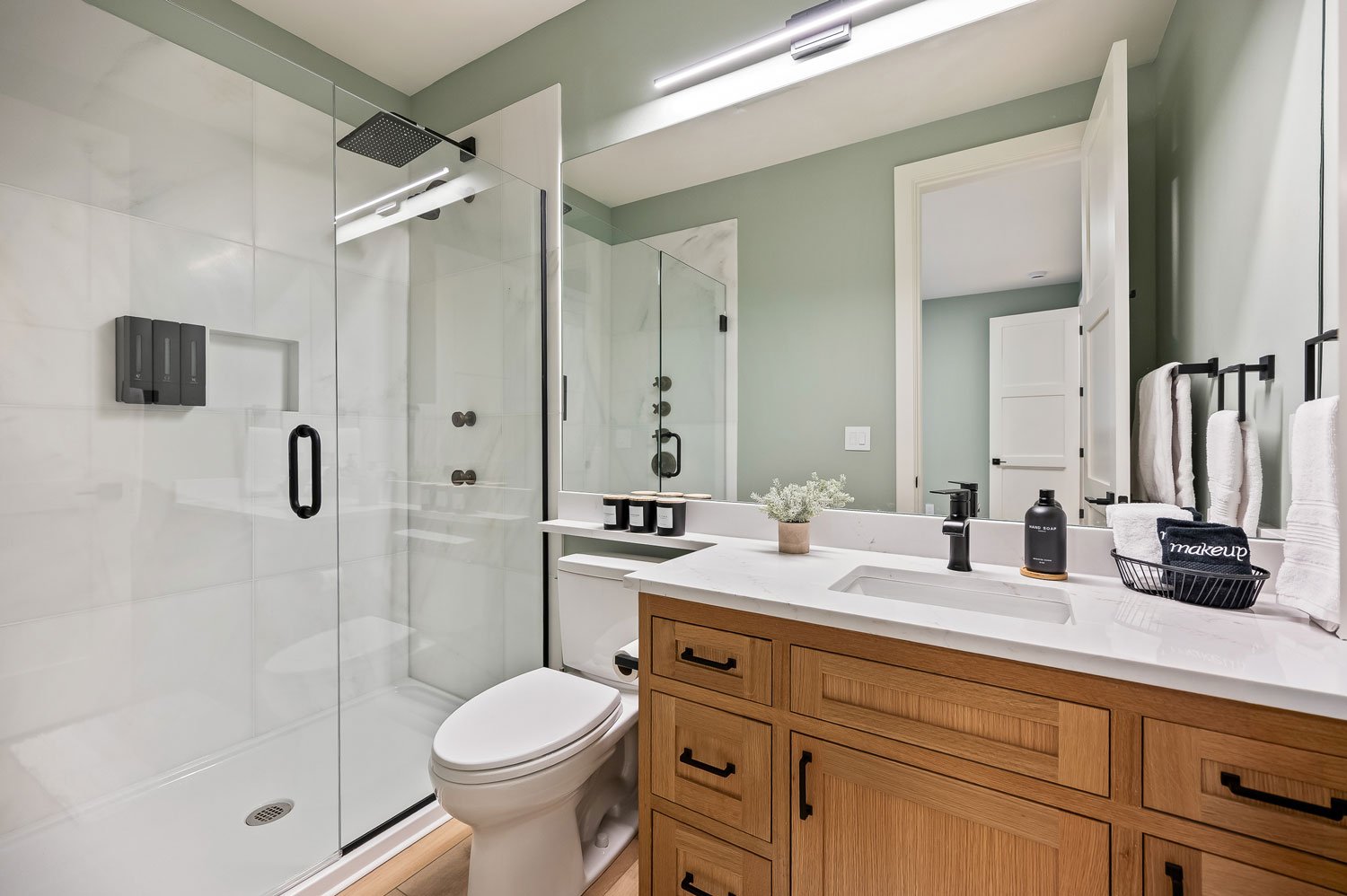 Modern bathroom with glass shower enclosure, white toilet, wooden vanity with black hardware, large mirror, black faucet, and black decor accents.