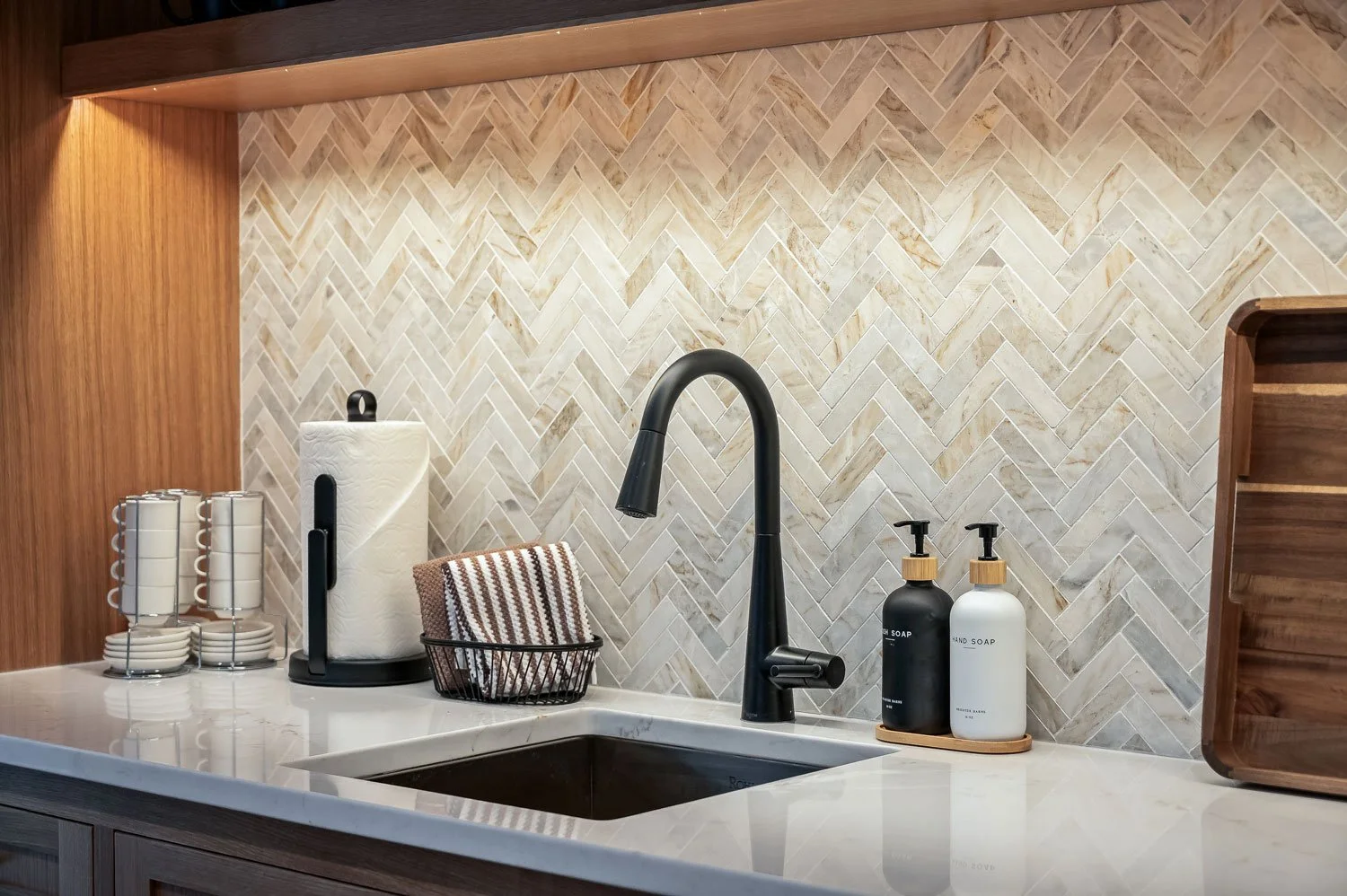 Kitchen countertop with a black faucet over a sink, paper towel holder, paper towels, dishes, and soap dispensers, with a herringbone tile backsplash.