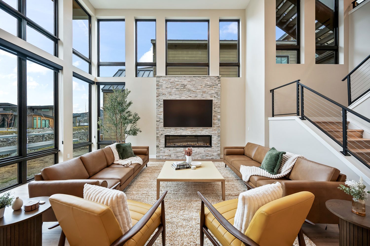 Modern living room with large windows, two brown leather couches, two yellow armchairs, a central beige coffee table, balcony stairs, a wall-mounted TV, and a stone fireplace.
