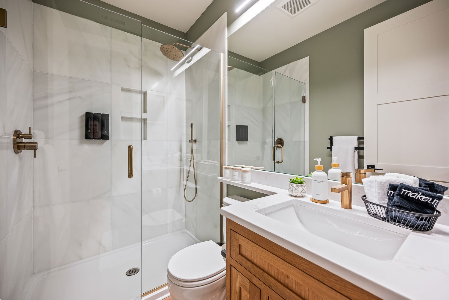 A modern bathroom with a glass-enclosed shower area, white marble walls, a white toilet, a wooden vanity with a white sink, a large mirror, and various toiletries including soap dispensers, a plant, and towels.