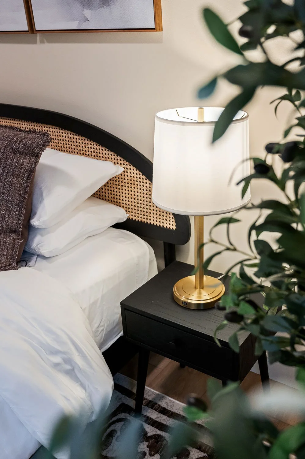 A bedroom with a bed, pillows, a black nightstand, a white lampshade with a brass base, a black picture frame, and a plant partially covering the view.