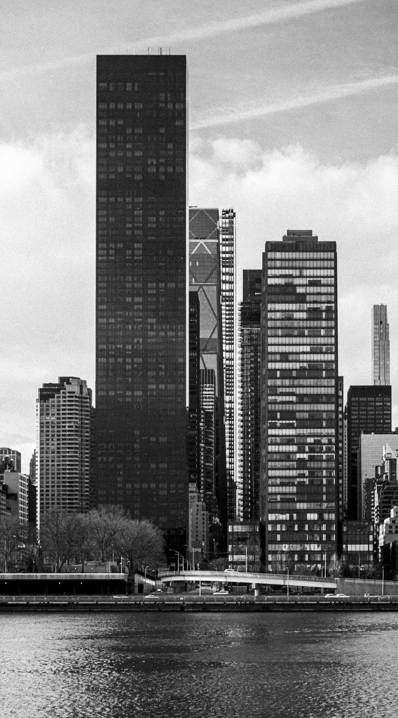 Black and white photo of modern city skyline with tall skyscrapers and a river in the foreground.
