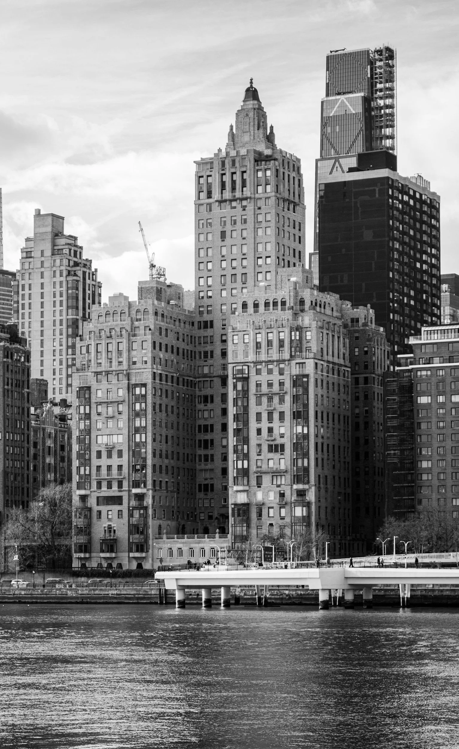 Black and white photo of a city skyline with tall buildings, a river in the foreground, and a bridge crossing the river.