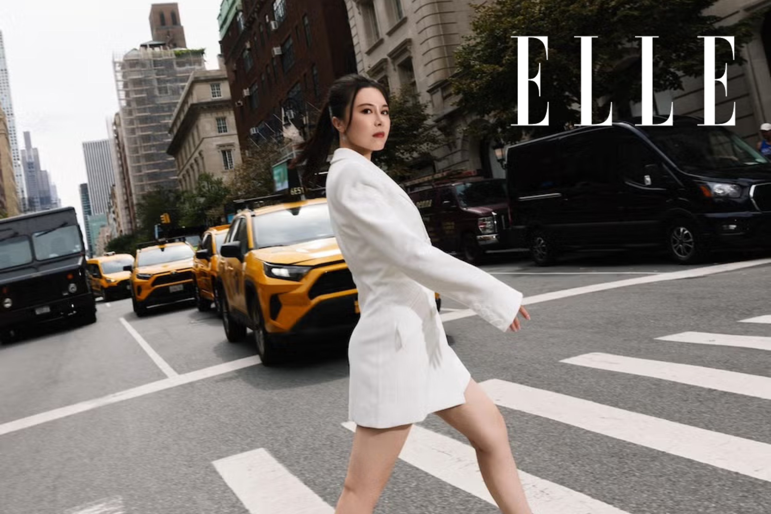 A woman walks across a city intersection with yellow taxis and black cars behind her, wearing a white blazer dress, with the word 'ELLE' in the top right corner.