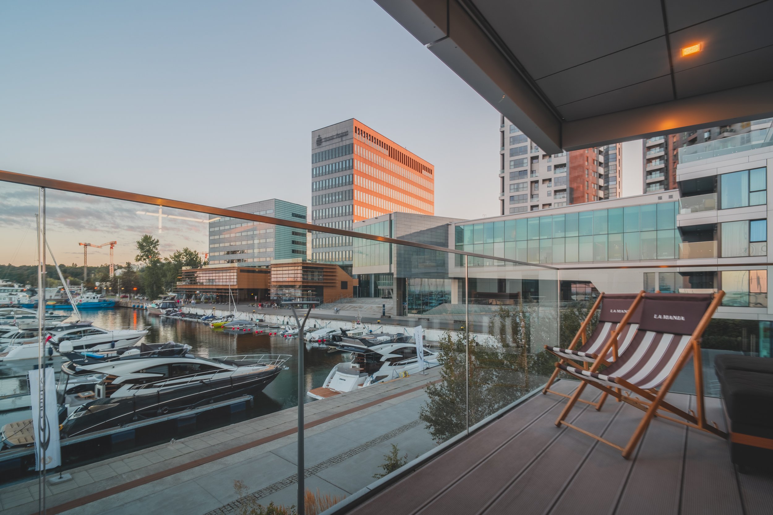 Balcony view of a marina with yachts, overlooking modern buildings during sunset.
