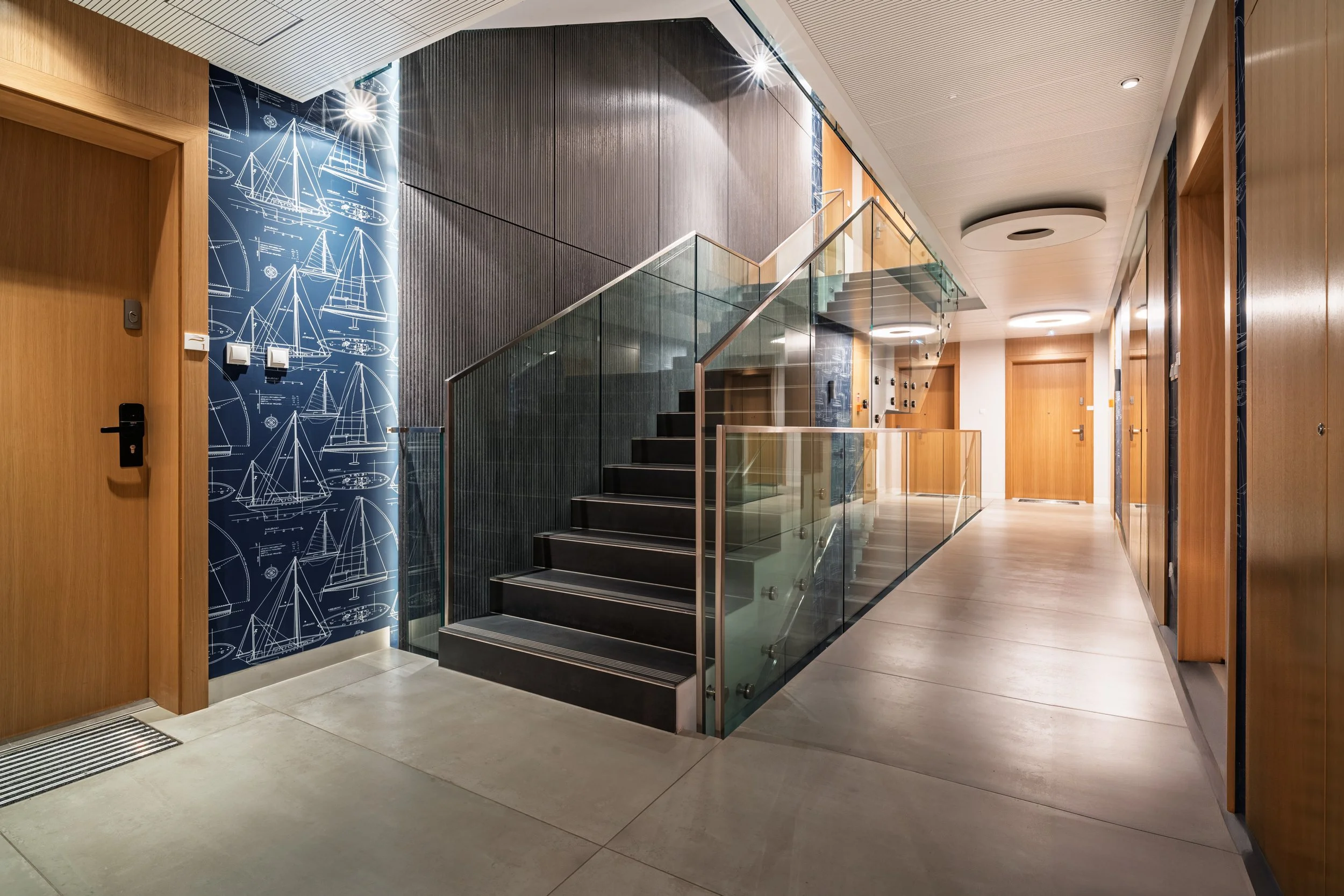 Modern residential building corridor with wooden doors, a staircase with glass railing, and nautical-themed blue wallpaper on one wall.