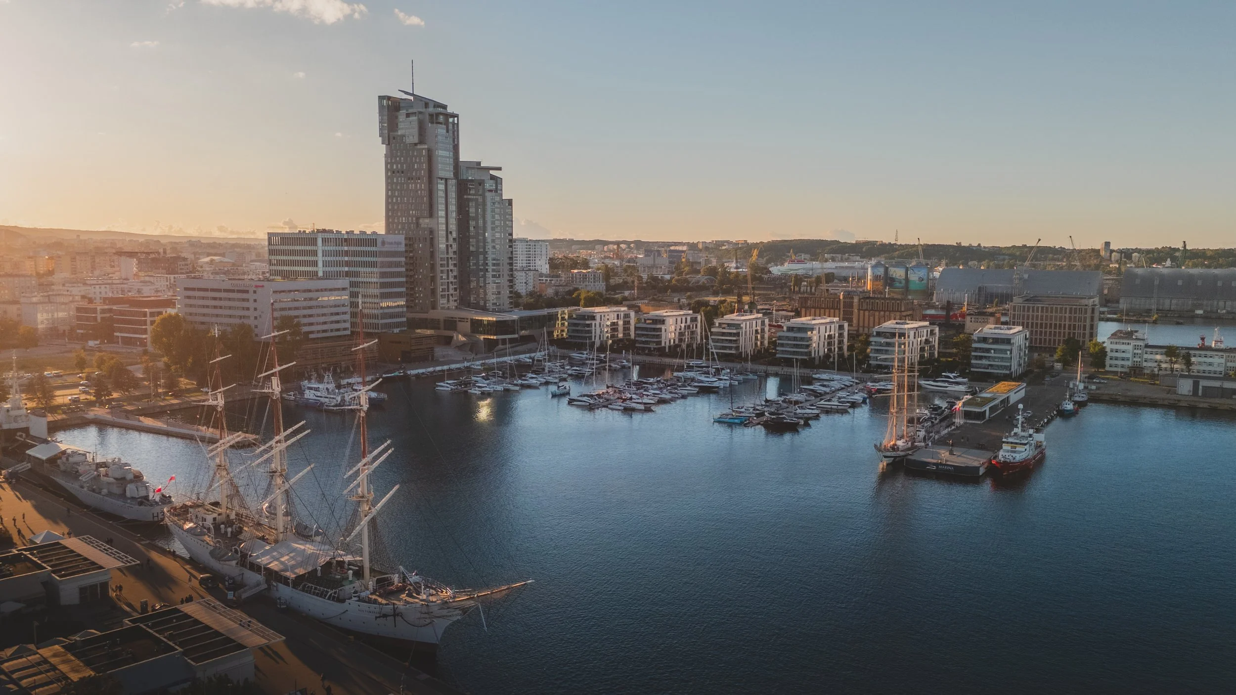 Aerial view of a marina with ships and boats docked, surrounded by modern high-rise buildings, during sunset.