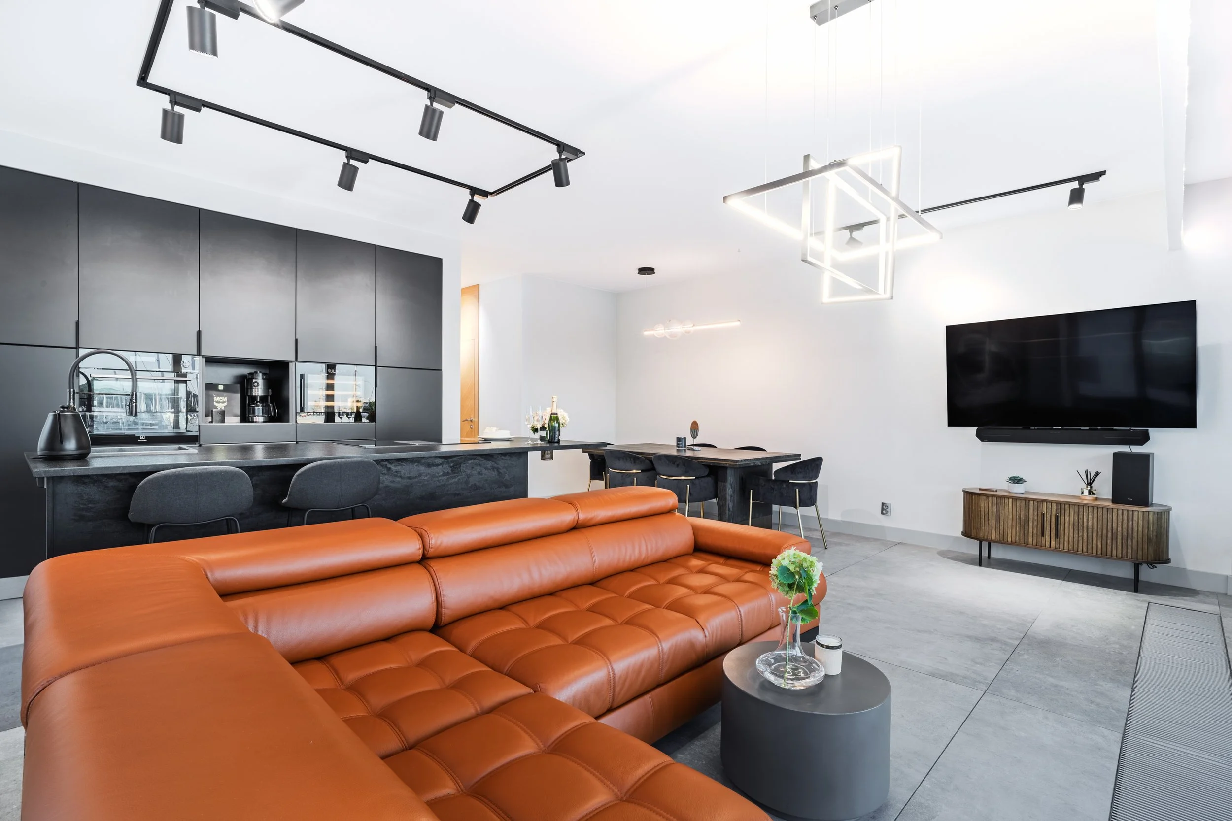 Modern open-concept living room and kitchen with a brown leather sectional sofa, black kitchen cabinets, bar stools, and a minimalist dining table. The room features contemporary lighting fixtures, a flat-screen TV, and light-colored walls and floors