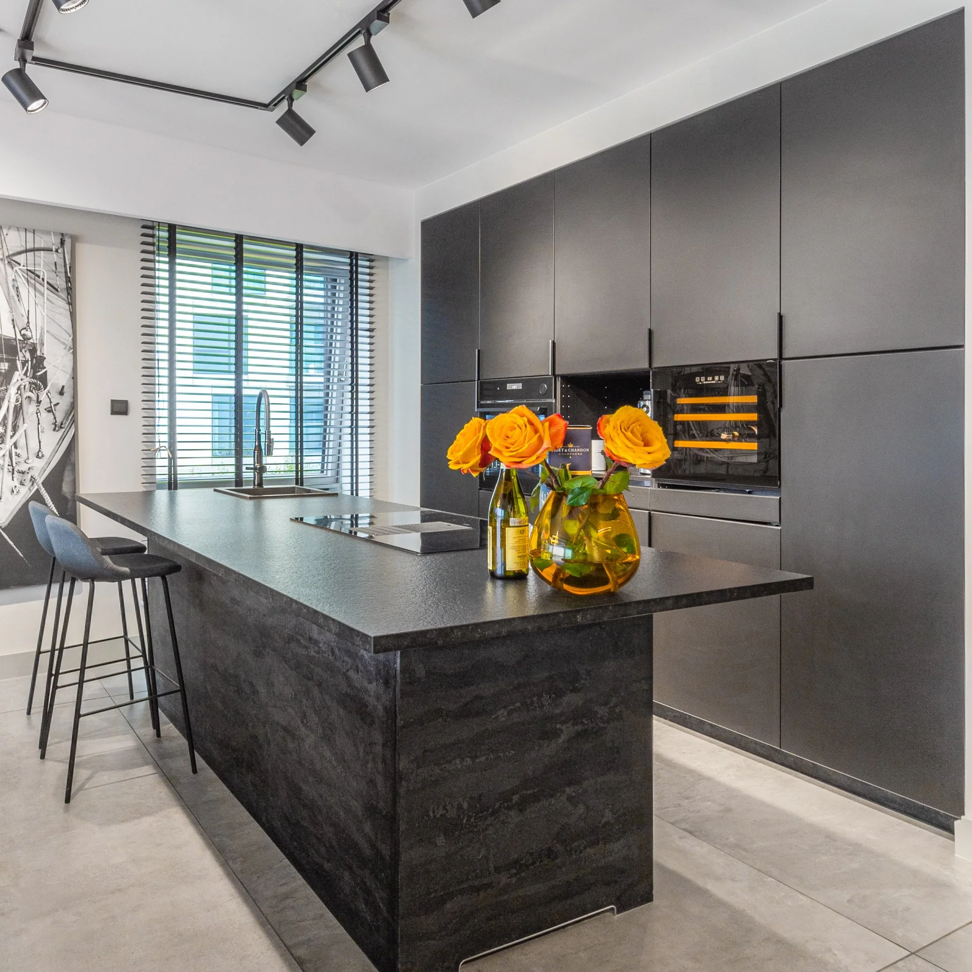 Modern kitchen with black cabinetry, a black island with a textured surface, and a sink beneath large window blinds. Two chairs are at the island, and a vase with orange roses is on the counter.