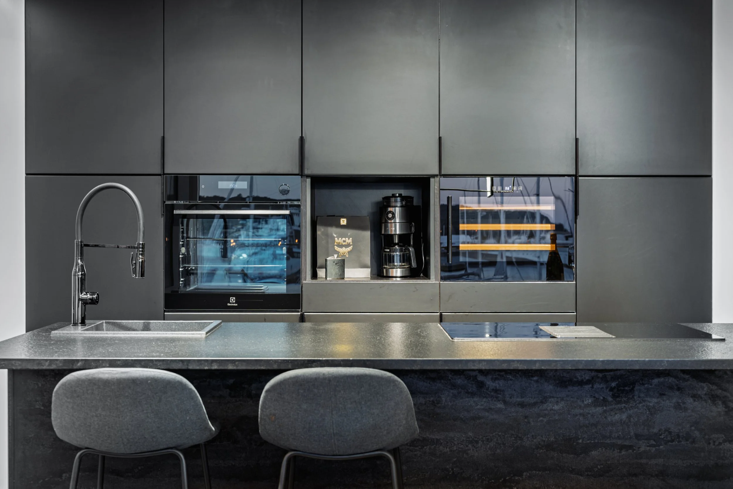 Modern kitchen with black cabinetry, built-in oven, coffee maker, and induction cooktop, gray countertop, and two gray chairs