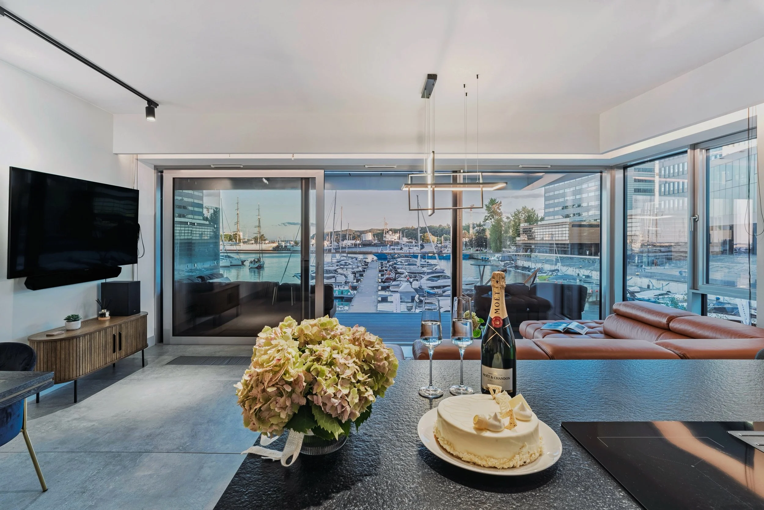 Modern living room with large windows overlooking a marina with boats, featuring a black countertop with a cake, champagne, flowers, and champagne glasses, and a wall-mounted TV.