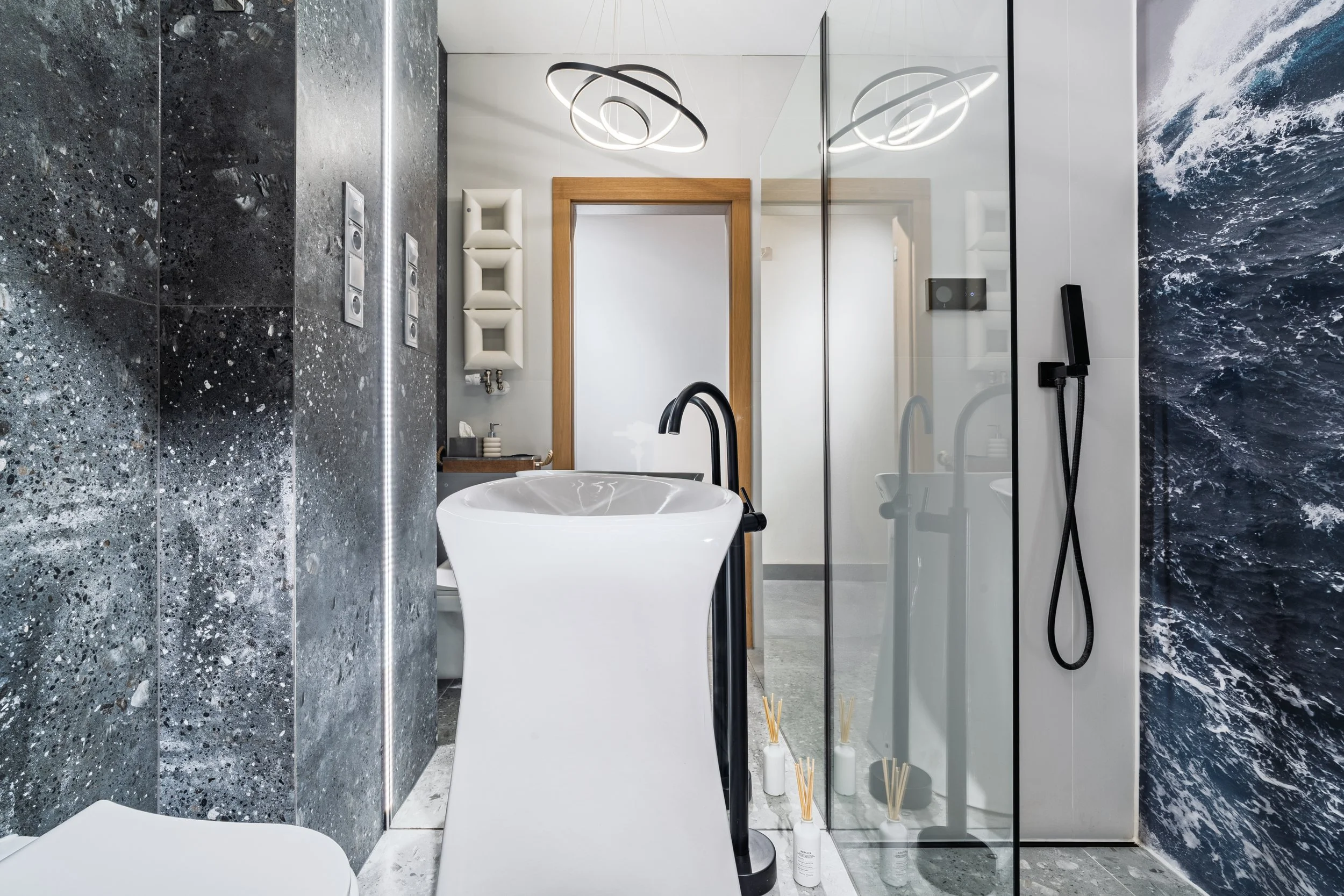 Modern bathroom with a freestanding white bathtub, black fixtures, a glass shower with a black handheld showerhead, and blue ocean-themed wall decor.