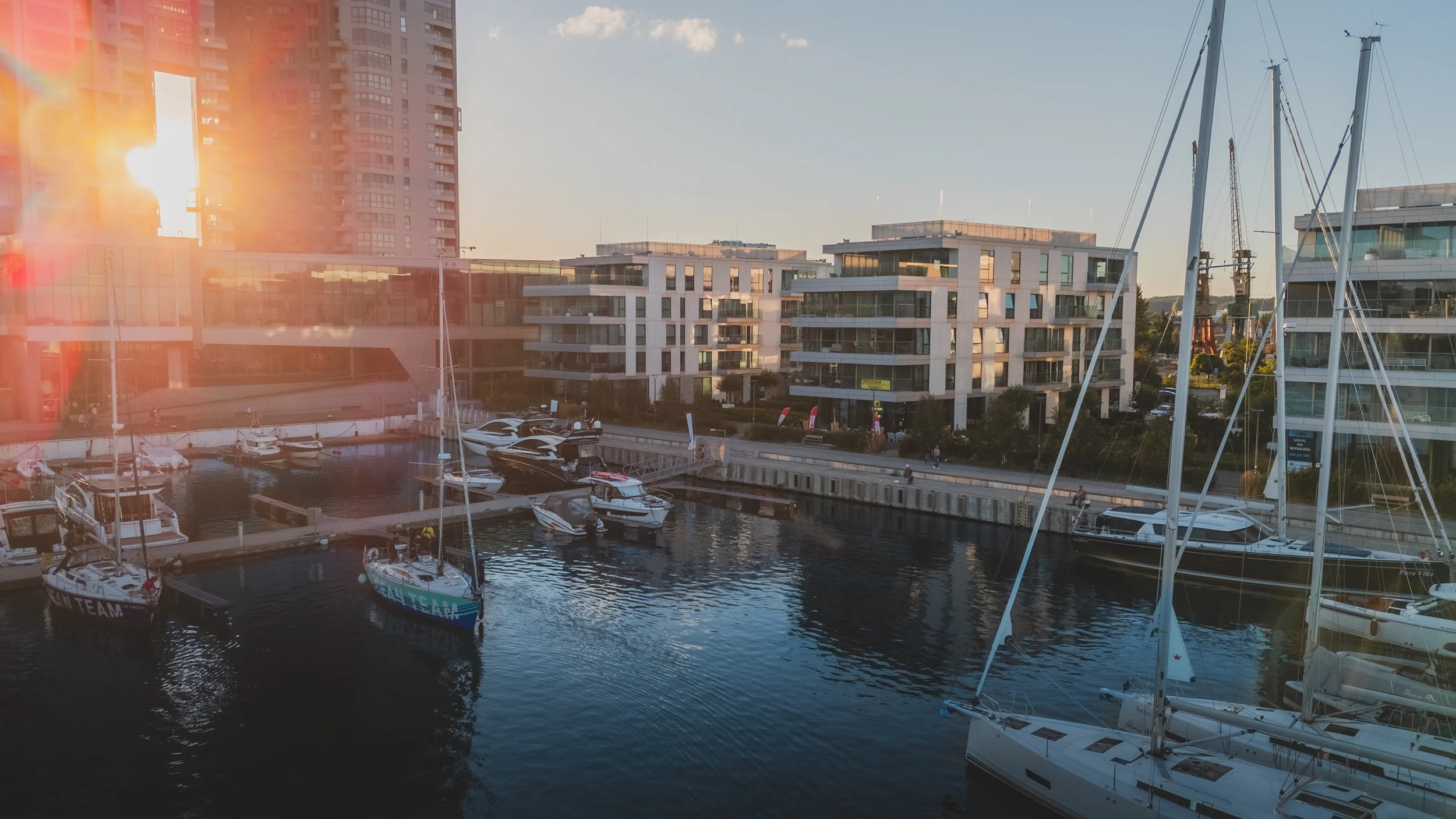 Sunset over a marina with boats docked and modern apartment buildings in the background.