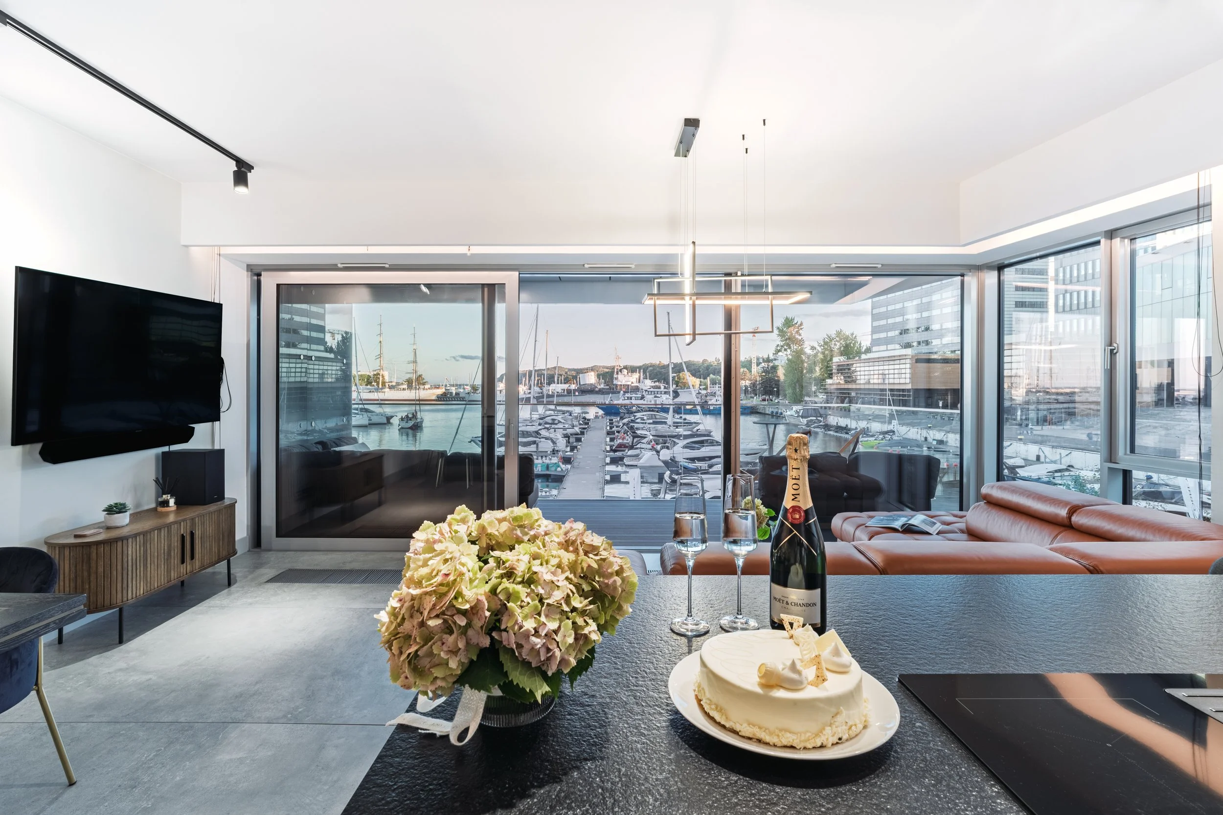 Modern living room with large windows overlooking a marina with boats, decorated for a celebration with a bottle of Moët & Chandon champagne, two glasses, a birthday cake, a bouquet of flowers, and a TV on the wall.