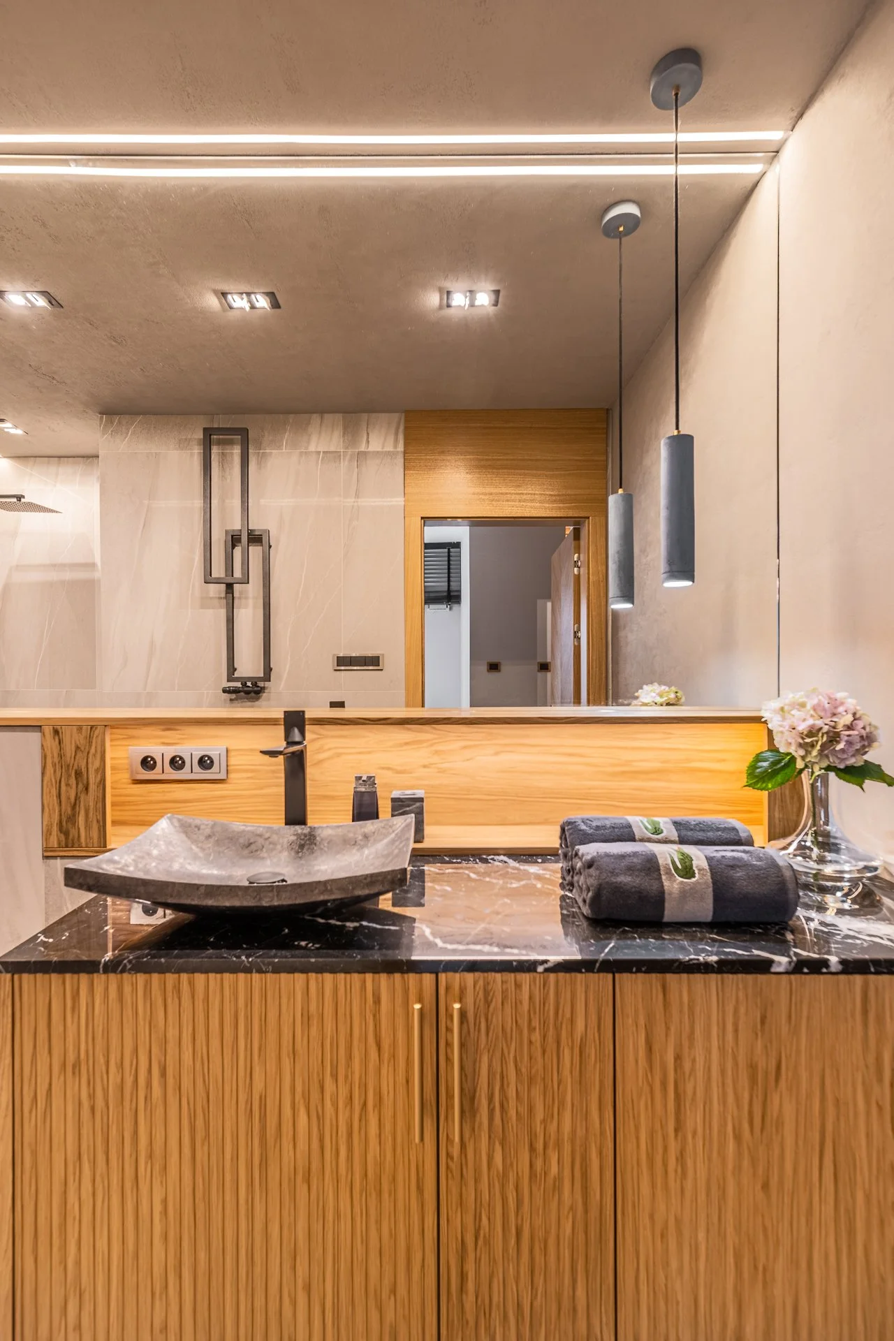 Modern bathroom with black marble countertop, stone vessel sink, black faucet, wooden cabinet, rolled gray towels, pink flowers, pendant lights, and large mirror.