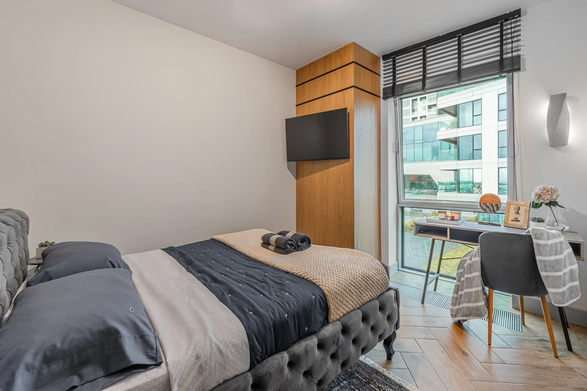 Contemporary bedroom with a gray upholstered bed, dark gray pillows, a beige blanket at the foot of the bed, a small side table, a wooden accent wall with a mounted flat-screen TV, a large window with a view of modern buildings, a gray chair covered with a striped towel, a small desk with decor, and a wall-mounted light fixture.