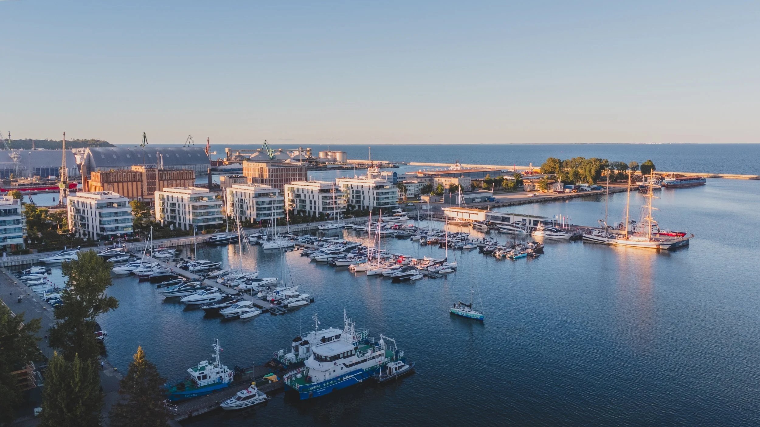 Aerial view of a marina with numerous boats and yachts docked, modern residential buildings nearby, industrial infrastructure in the background, and open water extending to the horizon.