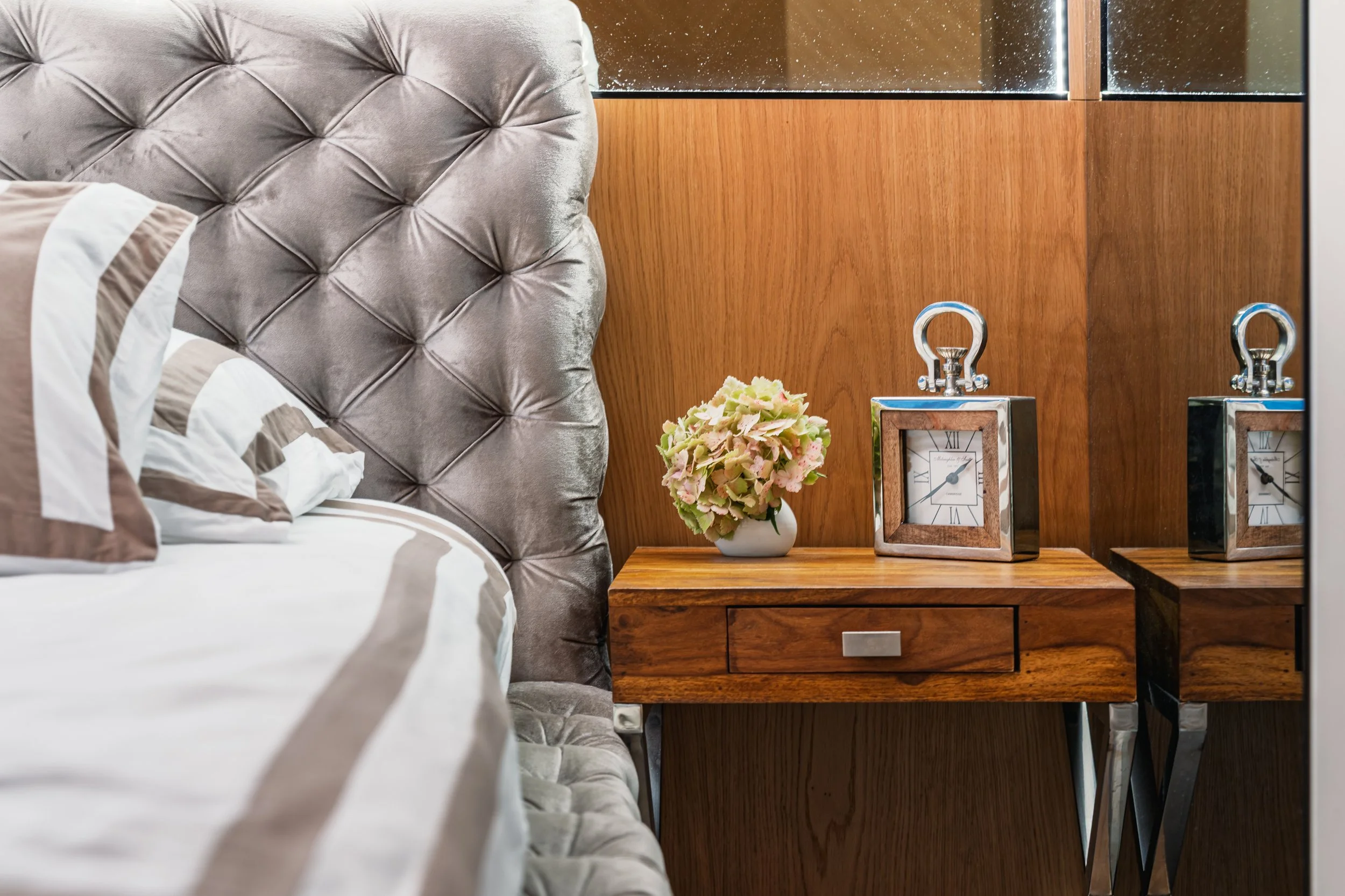 A bedroom corner featuring a tufted gray headboard, striped bed linens, a wooden nightstand with a drawer, a small white vase with pink and green flowers, and two silver-framed clocks on the nightstand. The background has wood wall paneling.