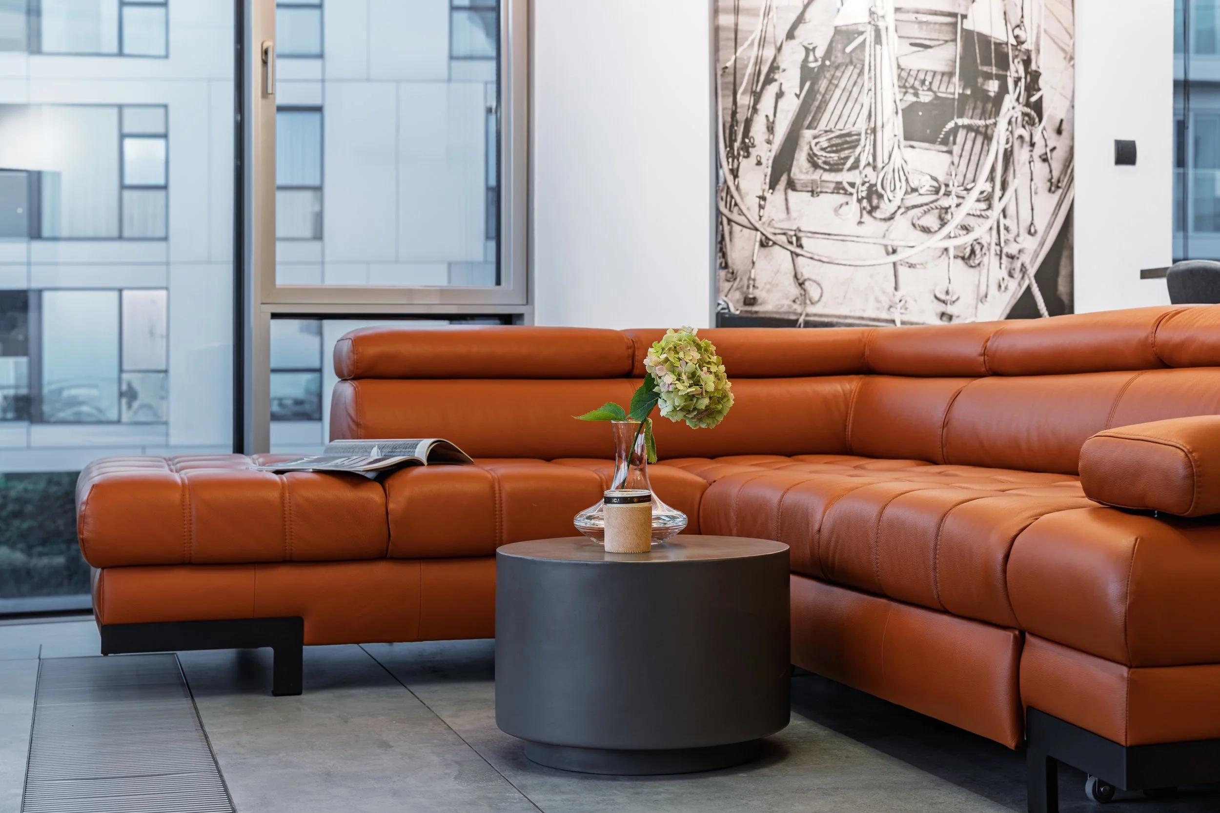 Modern office lounge with orange leather sectional sofa, a round black coffee table with a vase of green hydrangea, an open magazine, large windows, and a black-and-white abstract artwork on the wall.