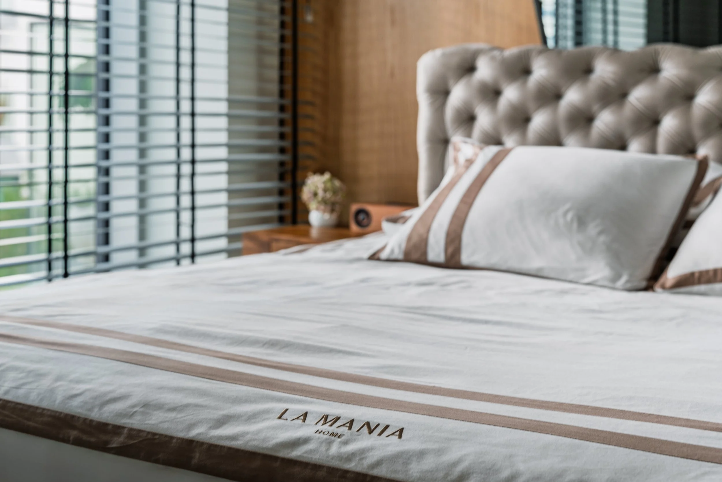 A neatly made bed with a tufted beige headboard, white and brown striped pillows, and a white blanket with beige and brown stripes, labeled 'LA MANIA HOME'; a wooden nightstand with a vase of flowers and a speaker, and a large window with horizontal 