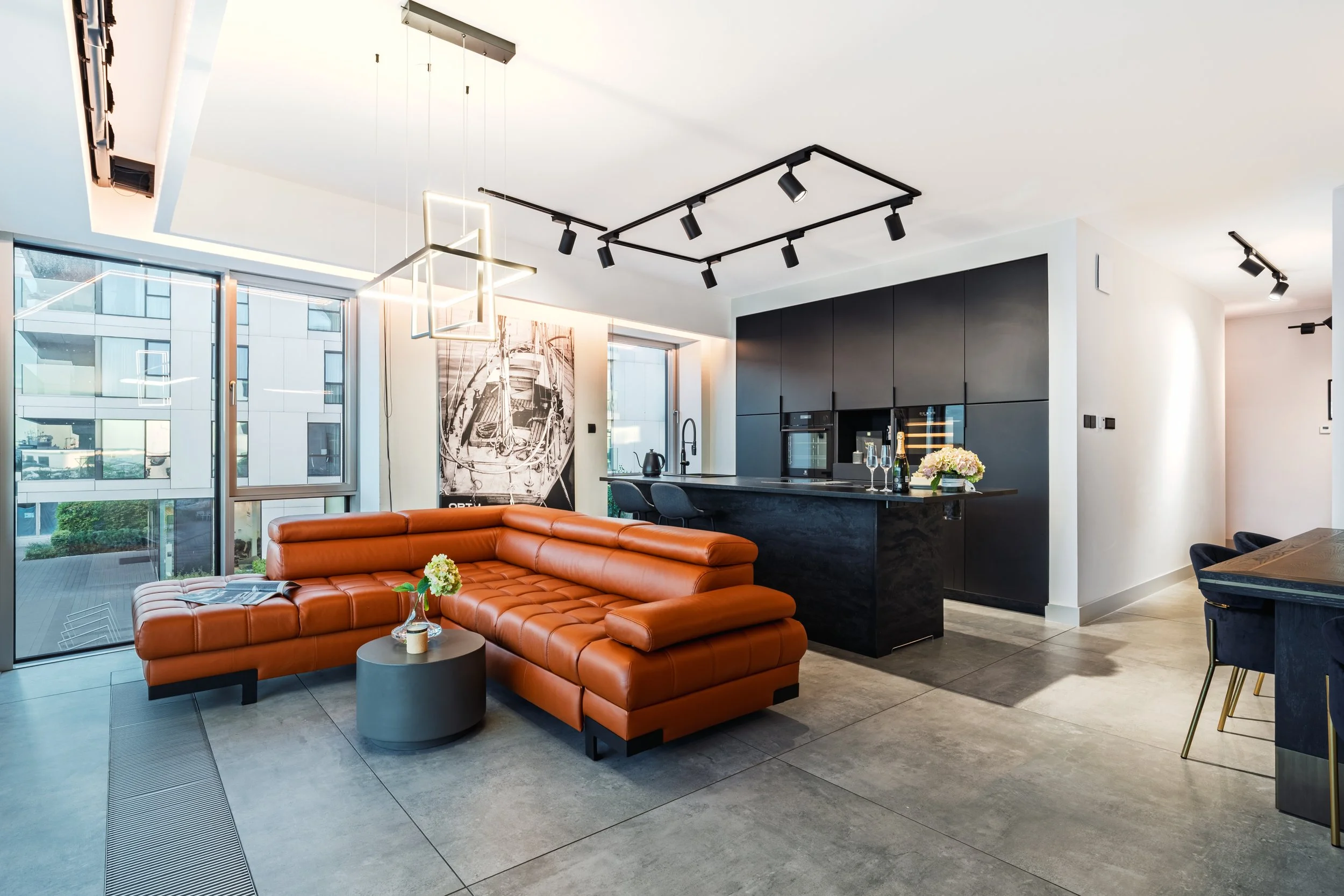 Modern living room with large windows, orange leather sectional sofa, round gray coffee table with a flower arrangement, black kitchen cabinetry, and artistic wall decor.